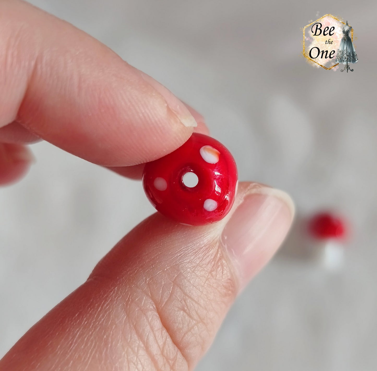 Perles en verre filé rouge et blanc en forme de champignon amanite tue-mouche à pois, percées verticalement, sur fond blanc. Gros plan trou du haut