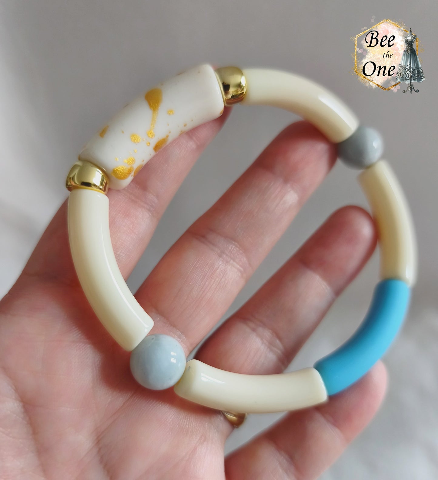 Set de trois bracelets élastiques faits main pour adulte, composés de perles tubes incurvées en acrylique aux teintes bleu azur, orange vif et crème, avec détails marbrés dorés et séparateurs dorés. Gros plan sur une perle blanche à marbrures dorées en acrylique