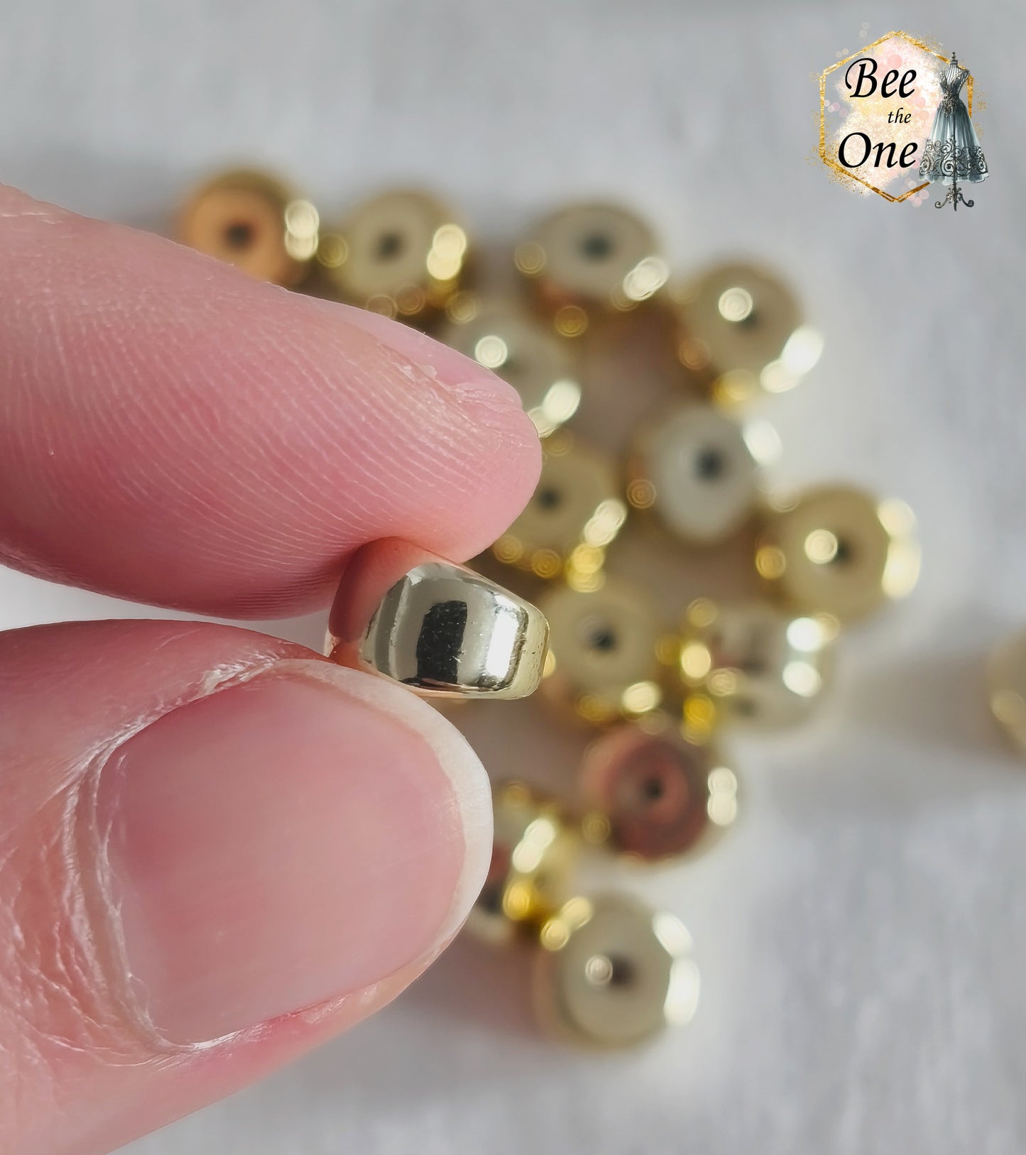 Perles tubes séparateurs incurvées en acrylique doré avec finition métallisée brillante et effet miroir, mesurant 11 mm, disposées sur un fond blanc texturé. Gros plan d'une perle entre deux doigts