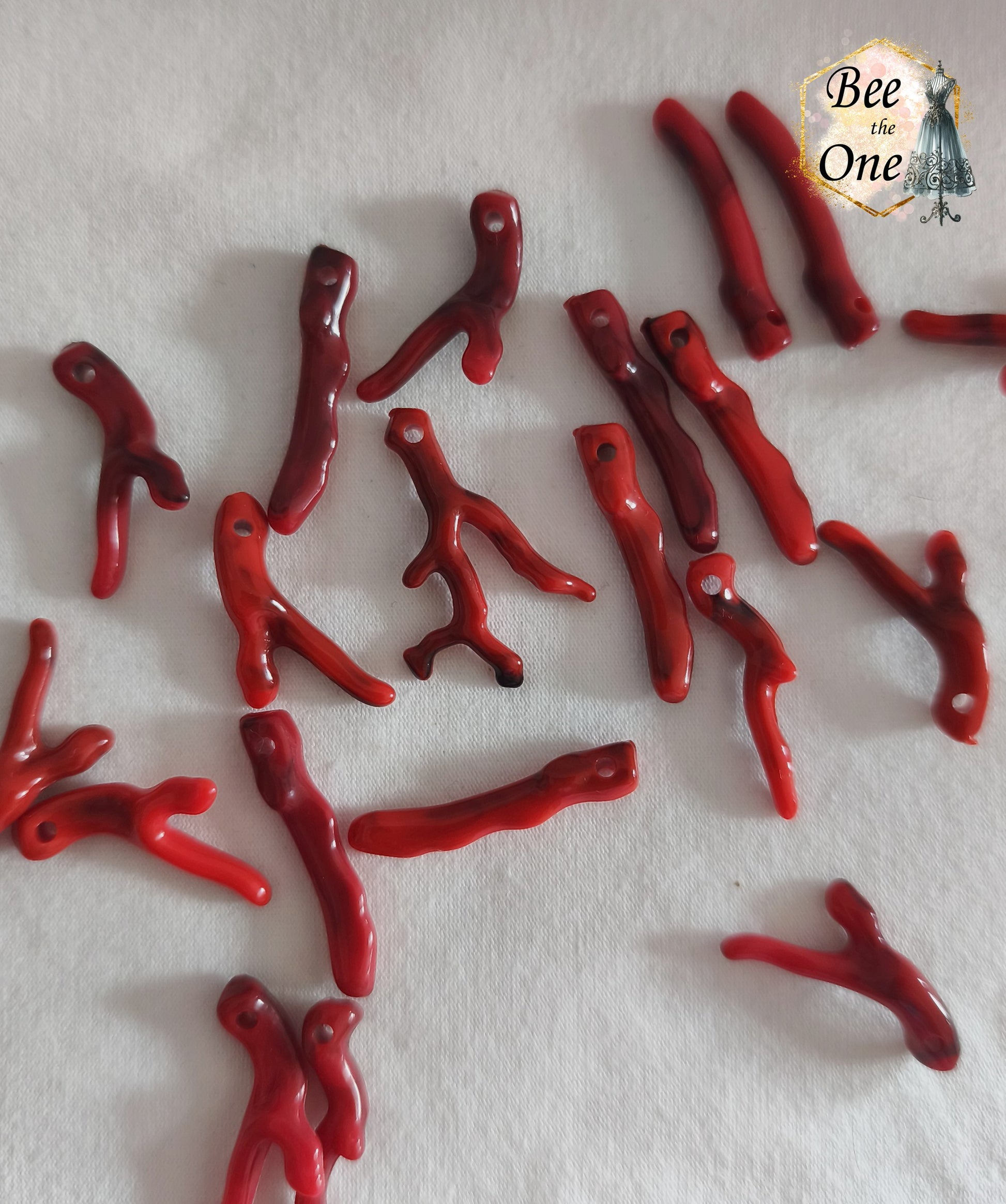 Breloques en acrylique imitation branches de corail rouge et bordeaux, formes organiques ramifiées et bâtons pré-percés d'environ 3 cm, posés sur fond blanc texturé.