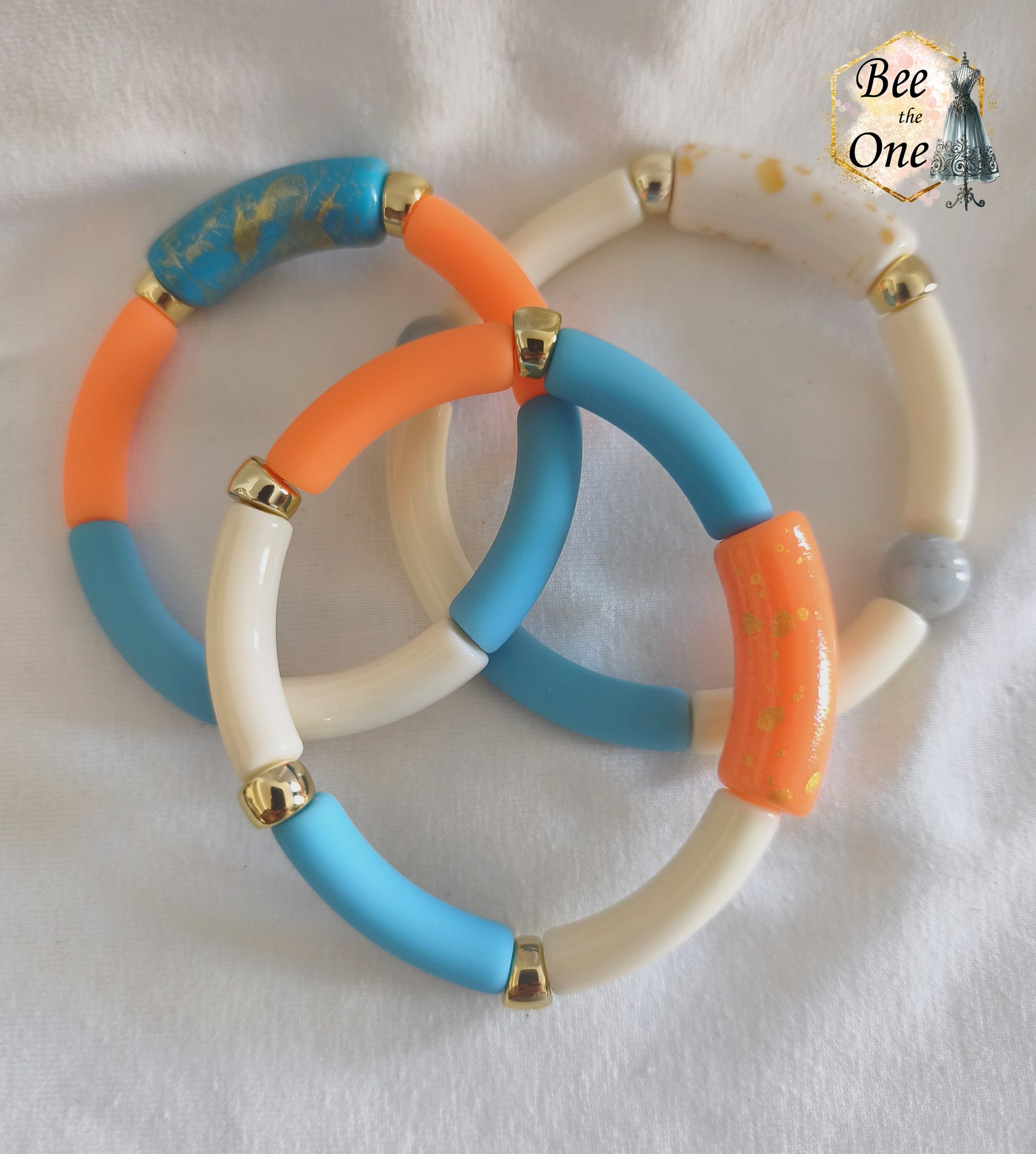 Set de trois bracelets élastiques faits main pour adulte, composés de perles tubes incurvées en acrylique aux teintes bleu azur, orange vif et crème, avec détails marbrés dorés et séparateurs dorés, présentés sur un fond blanc.