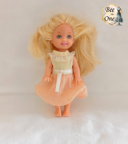 Petite robe de princesse pour poupée Kelly (Shelly), issue du film Barbie et le Cheval Magique (2005). Le haut est blanc scintillant avec un col volanté blanc, la jupe est en tulle pêche, portée par une petite poupée blonde sur fond blanc.