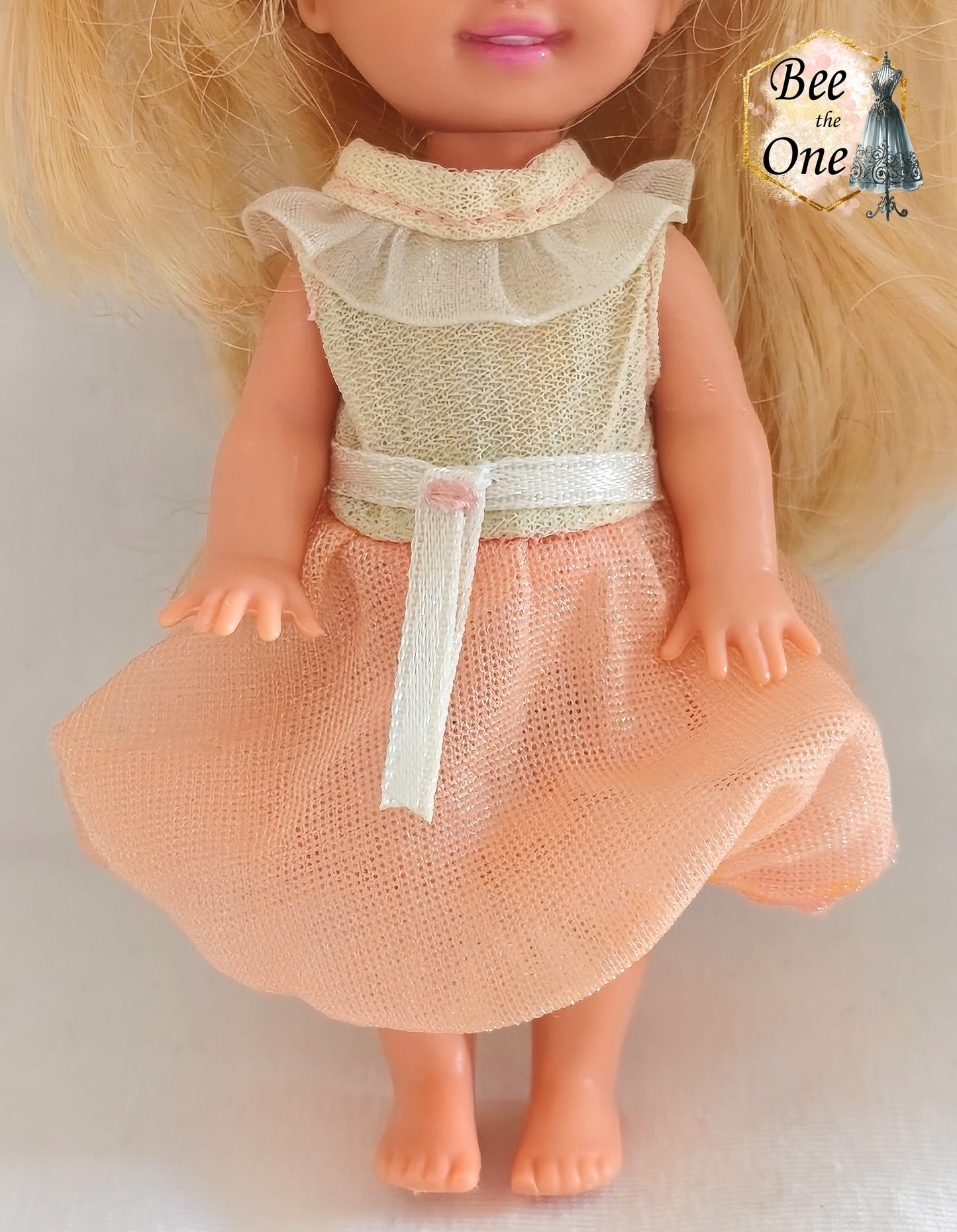 Petite robe de princesse pour poupée Kelly (Shelly), issue du film Barbie et le Cheval Magique (2005). Le haut est blanc scintillant avec un col volanté blanc, la jupe est en tulle pêche, portée par une petite poupée blonde sur fond blanc. Gros plan devant