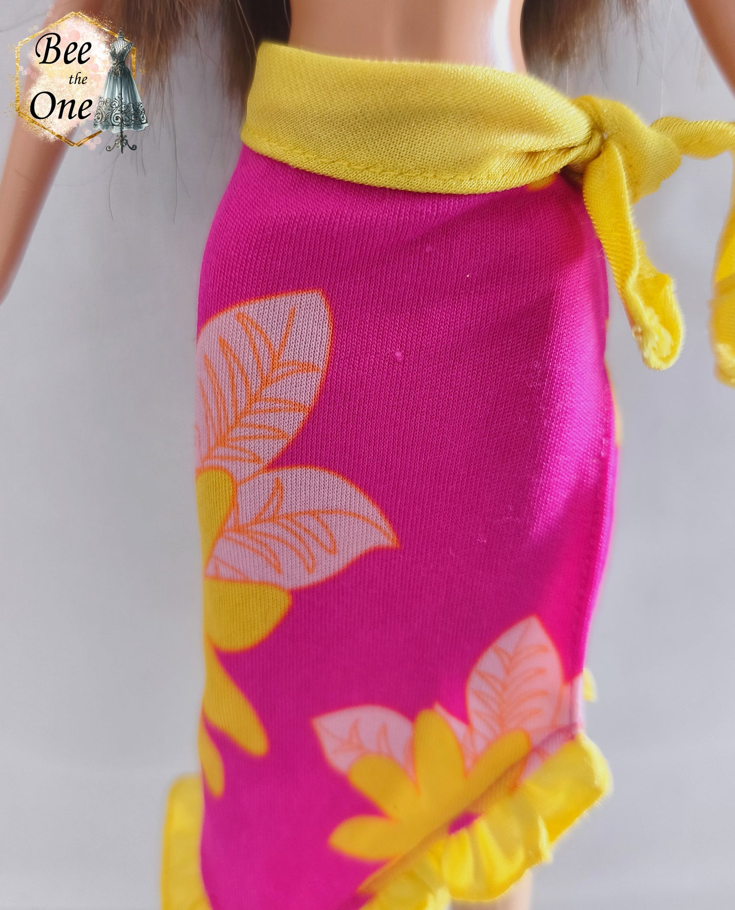 Paréo sarong de plage rose fuchsia à motifs de fleurs tropicales jaunes et orange avec bordure et ceinture nouée jaune fluo, vêtement d'origine pour poupée Barbie Tropical Holiday, porté par une poupée brune sur fond blanc. Gros plan devant