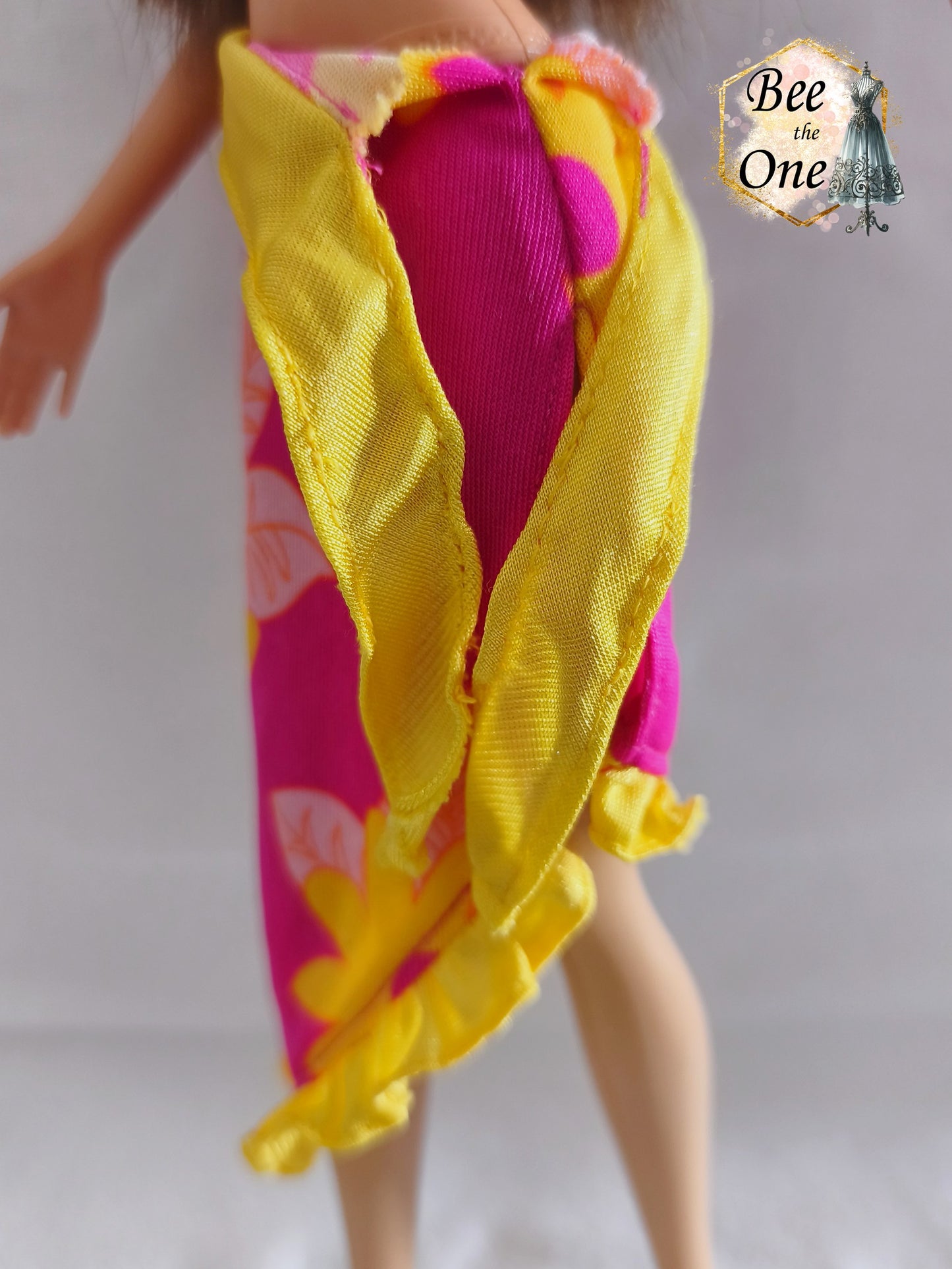 Paréo sarong de plage rose fuchsia à motifs de fleurs tropicales jaunes et orange avec bordure et ceinture nouée jaune fluo, vêtement d'origine pour poupée Barbie Tropical Holiday, porté par une poupée brune sur fond blanc. Gros plan ceinture