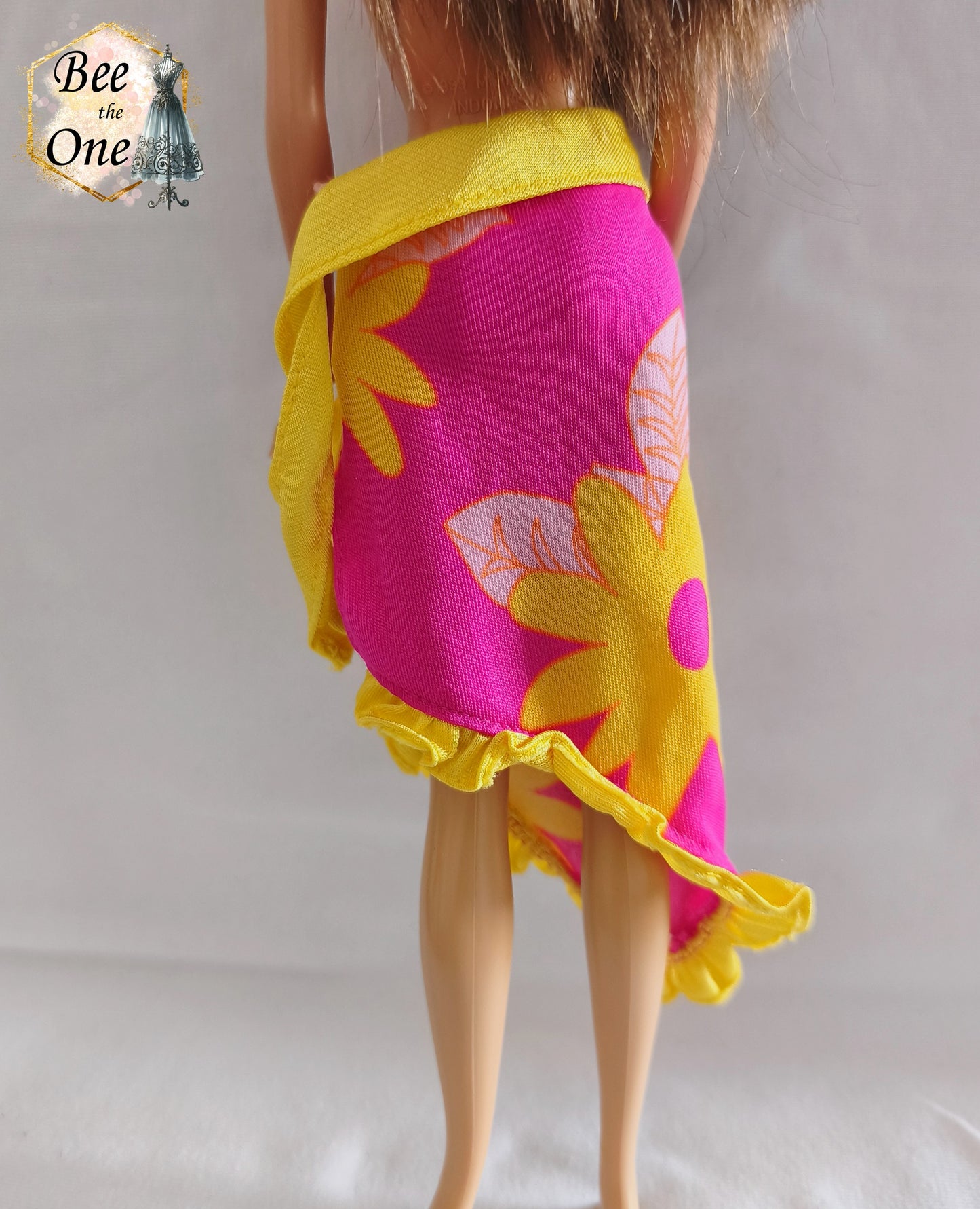 Paréo sarong de plage rose fuchsia à motifs de fleurs tropicales jaunes et orange avec bordure et ceinture nouée jaune fluo, vêtement d'origine pour poupée Barbie Tropical Holiday, porté par une poupée brune sur fond blanc. Gros plan dos