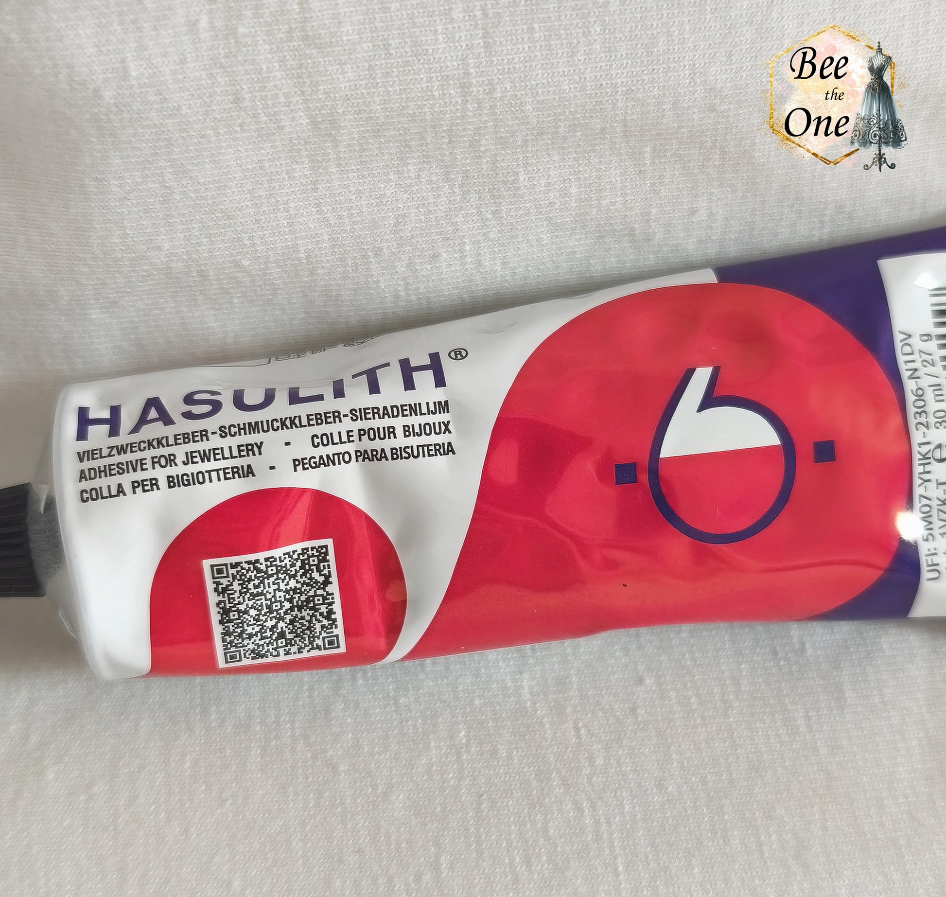 Un tube de colle Hasulith classique de 30 ml (27g) dans son emballage d'originerouge, violet et blanc, posé sur un fond blanc texturé. Gros plan devant