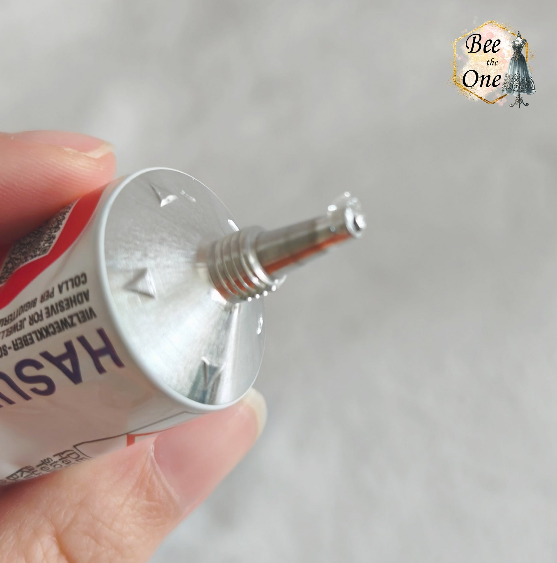 Un tube de colle Hasulith classique de 30 ml (27g) dans son emballage d'originerouge, violet et blanc, posé sur un fond blanc texturé. Gros plan embout