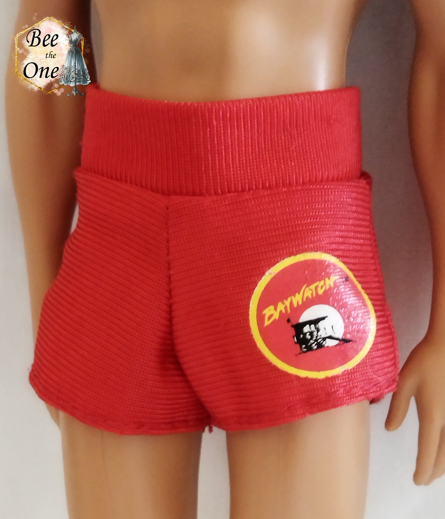 Short de plage pour poupée Ken, rouge avec un patch Baywatch sur le côté. Gros plan vue devant