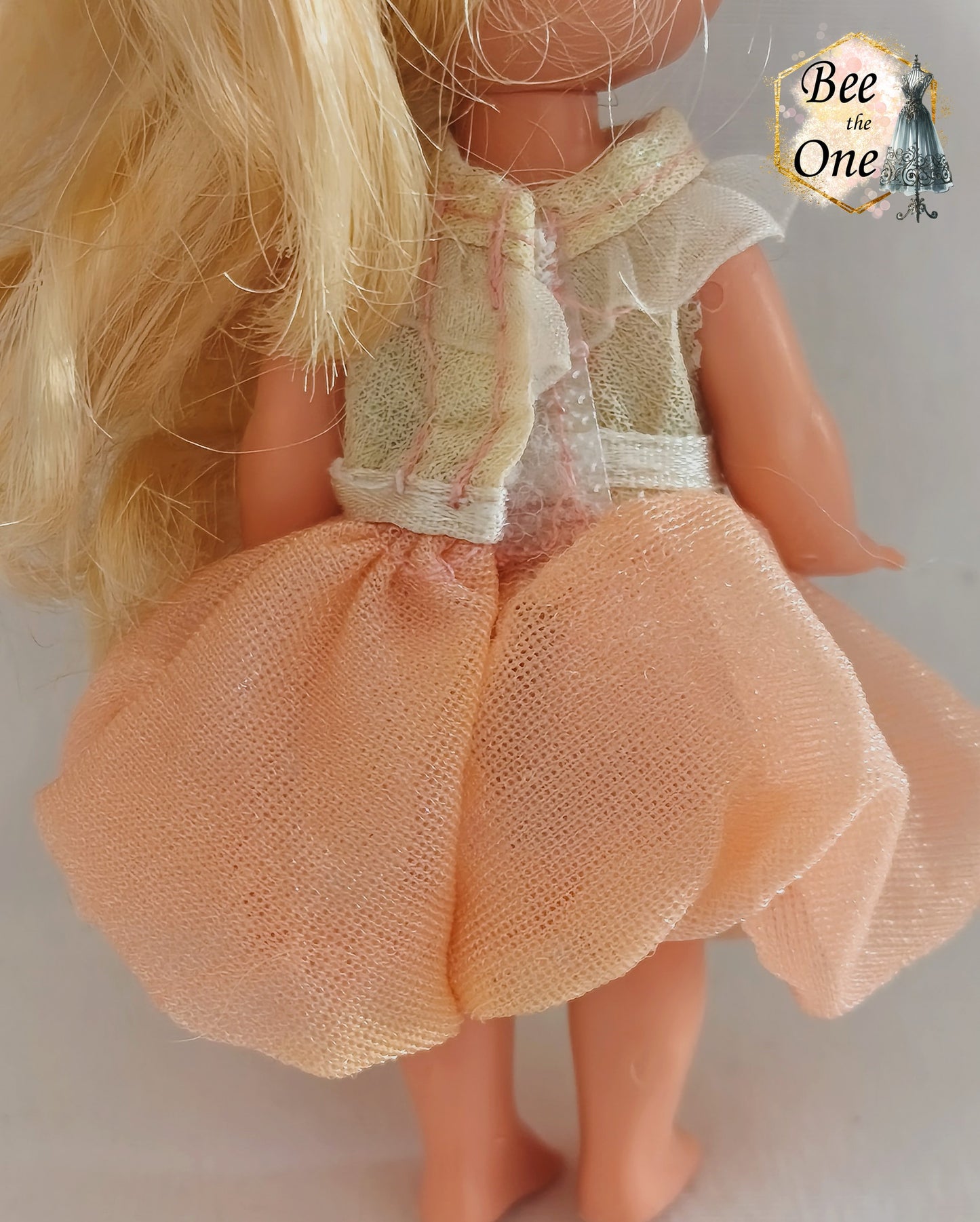 Petite robe de princesse pour poupée Kelly (Shelly), issue du film Barbie et le Cheval Magique (2005). Le haut est blanc scintillant avec un col volanté blanc, la jupe est en tulle pêche, portée par une petite poupée blonde sur fond blanc. Gros plan dos