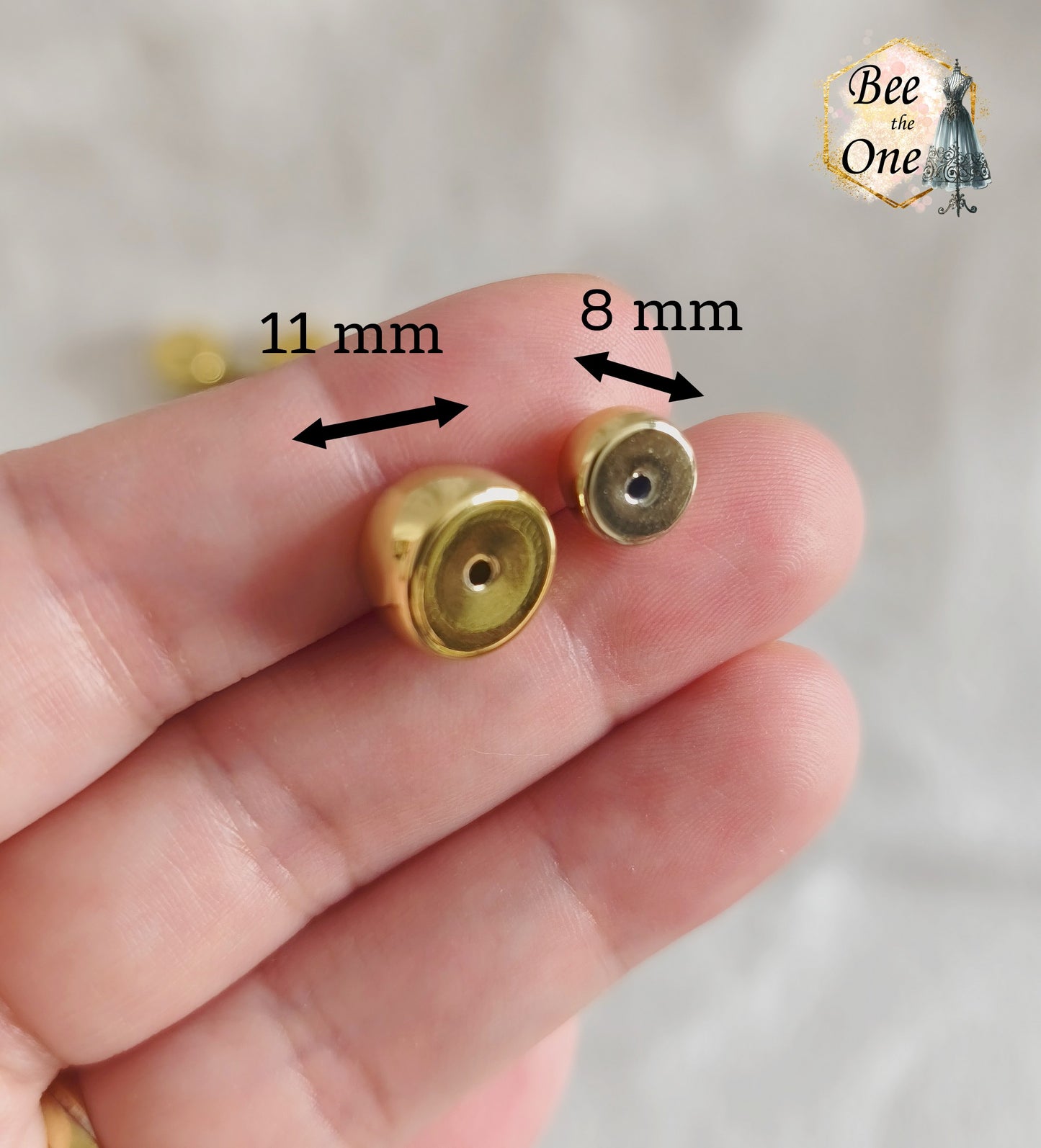 Perles tubes séparateurs incurvées en acrylique doré avec finition métallisée brillante et effet miroir, mesurant 11 mm, disposées sur un fond blanc texturé. Gros plan d'une perle entre deux doigts. Gros plan d'une perle de diamètre 11 mm et d'une autre identique en diamètre 8 mm