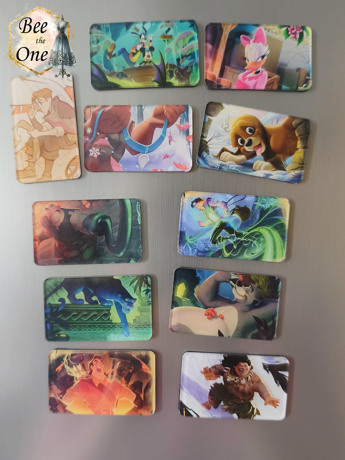 Série de onze magnets rectangulaires décoratifs illustrant des scènes et personnages fantastiques issus de cartes à collectionner TCG : Mickey et Dingo, Daisy, Milo Thatch, Bagheera, Baloo, Gaston et Maui, disposés sur une surface métallique grise.