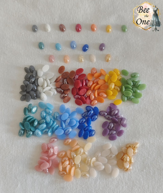Mini cabochons ovales lisses de 6x4 mm en acrylique, triés par petits tas de couleurs multicolores (bleu, vert, rouge, rose, orange, jaune, violet, blanc, gris, marron), certains mats et d'autres irisés, posés sur un fond blanc.