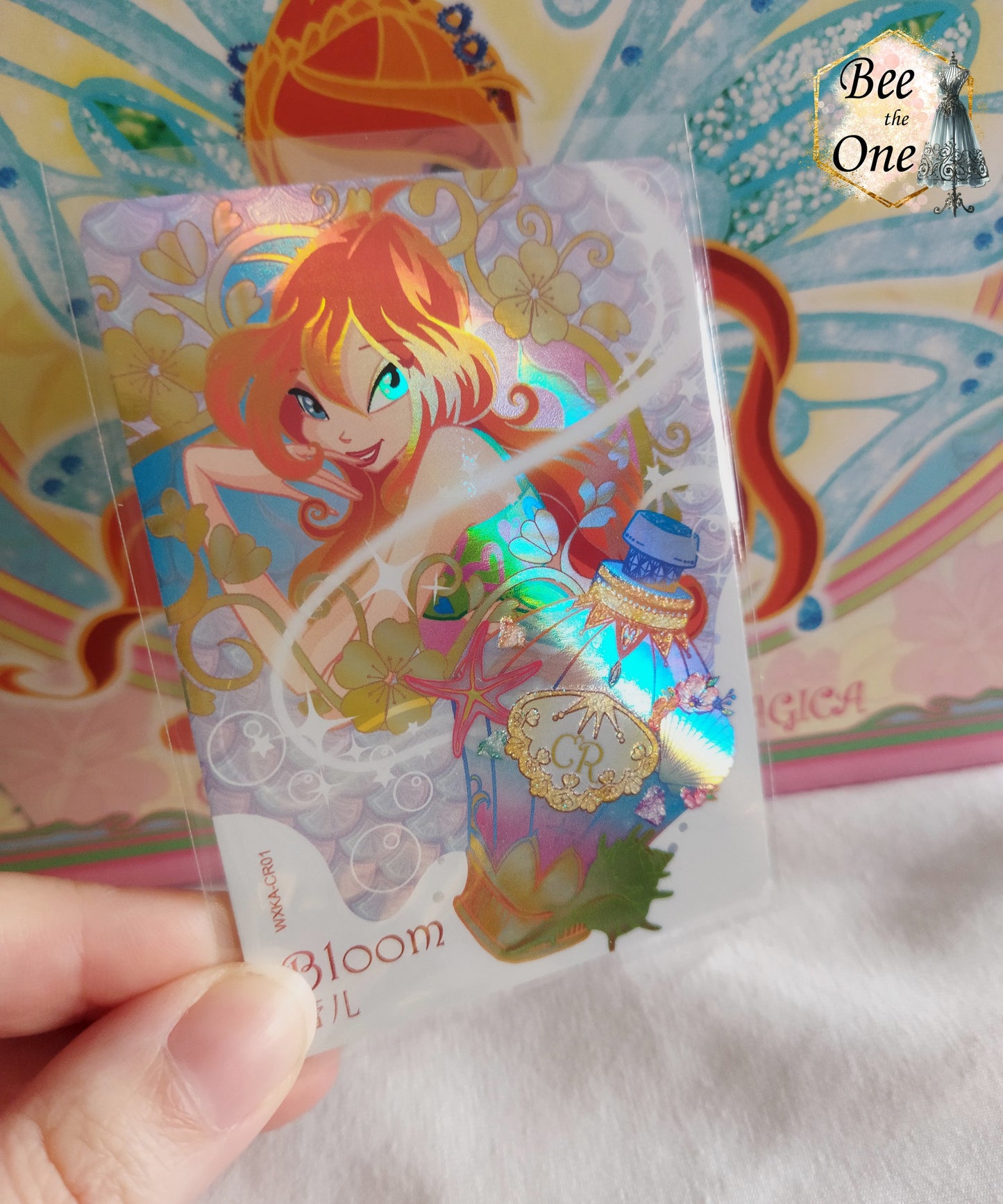 Cartes à collectionner inspirées de l'univers magique de Winx Club, mettant en scène Bloom dans un style coloré et féerique. La carte est ornée de motifs floraux, potion et de détails étincelants, évoquant la magie et l'aventure.