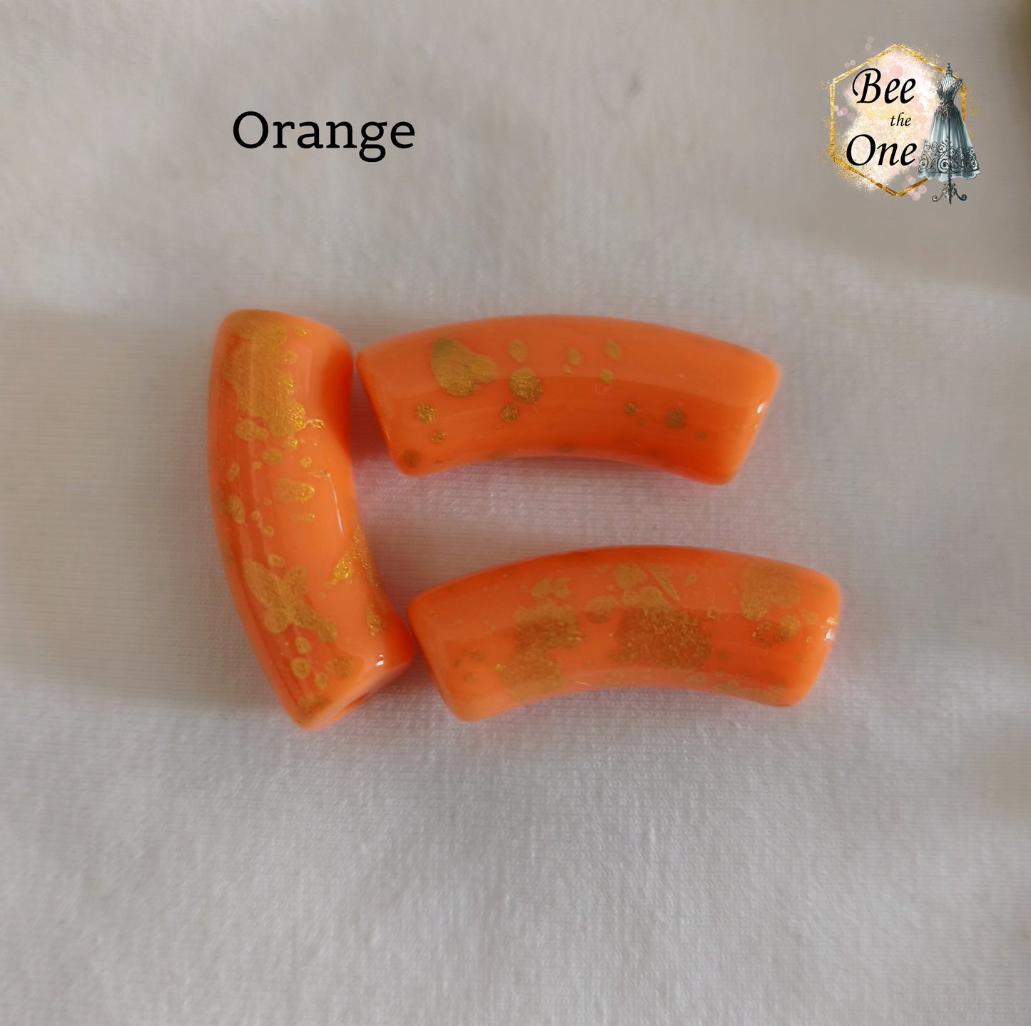 Grandes perles tubes incurvées en acrylique mesurant 34x11 mm. Les perles sont couleur orange et ornées d'éclats et de mouchetures dorées, disposées sur un fond blanc.