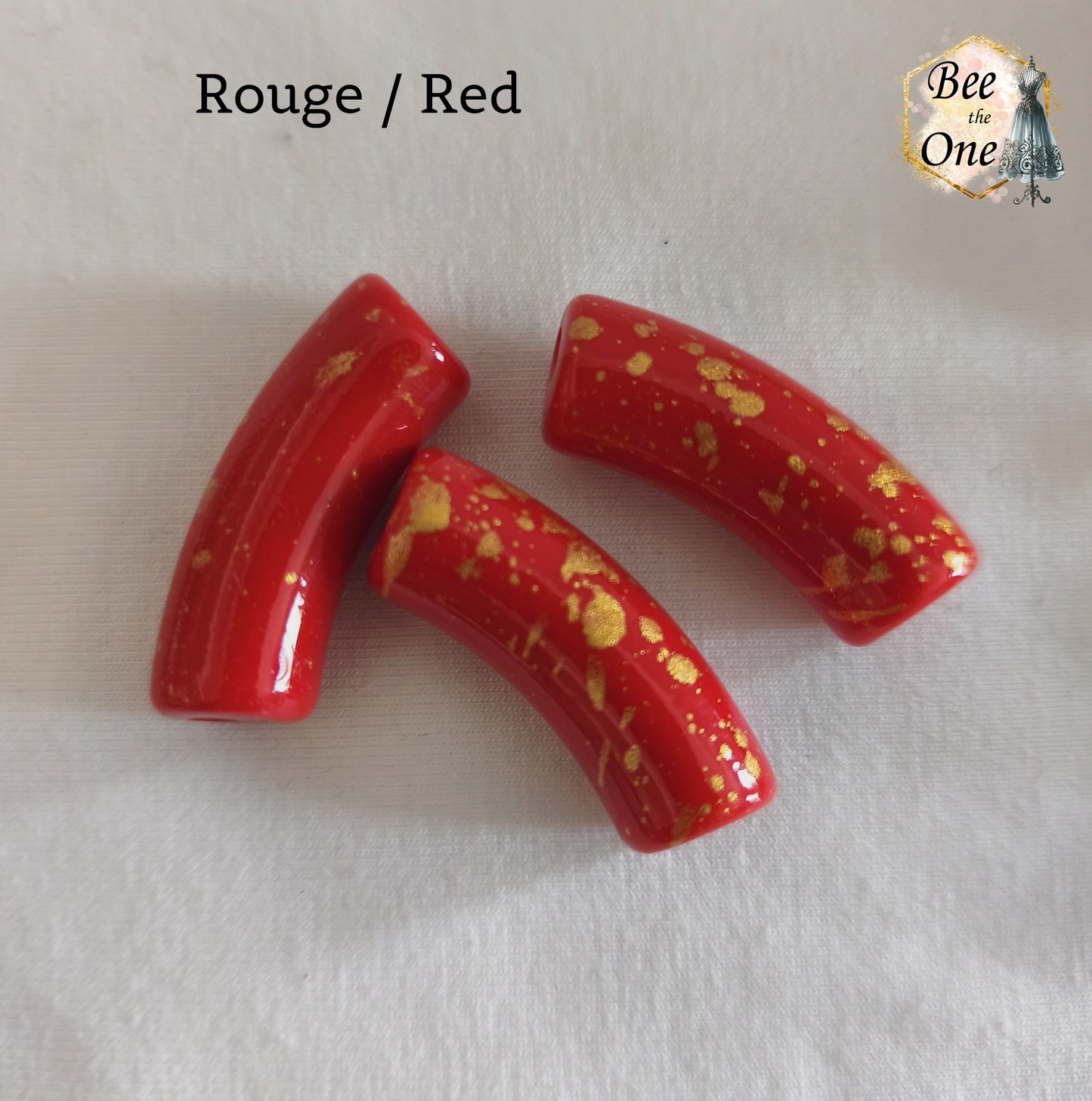 Grandes perles tubes incurvées en acrylique mesurant 34x11 mm. Les perles sont couleur rouge et ornées d'éclats et de mouchetures dorées, disposées sur un fond blanc.