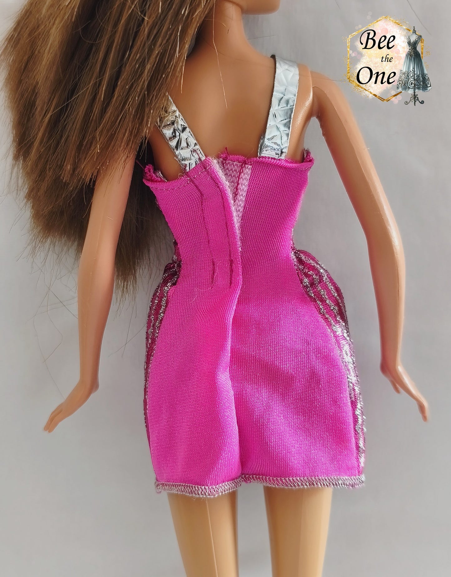 Robe courte de fête rose fuchsia brillante avec rayures verticales pailletées argentées, bretelles métallisées et découpes géométriques en losanges à la taille, vêtement d'origine pour poupée Barbie Fashionistas 2013, portée par une poupée brune sur fond blanc. Gros plan dos