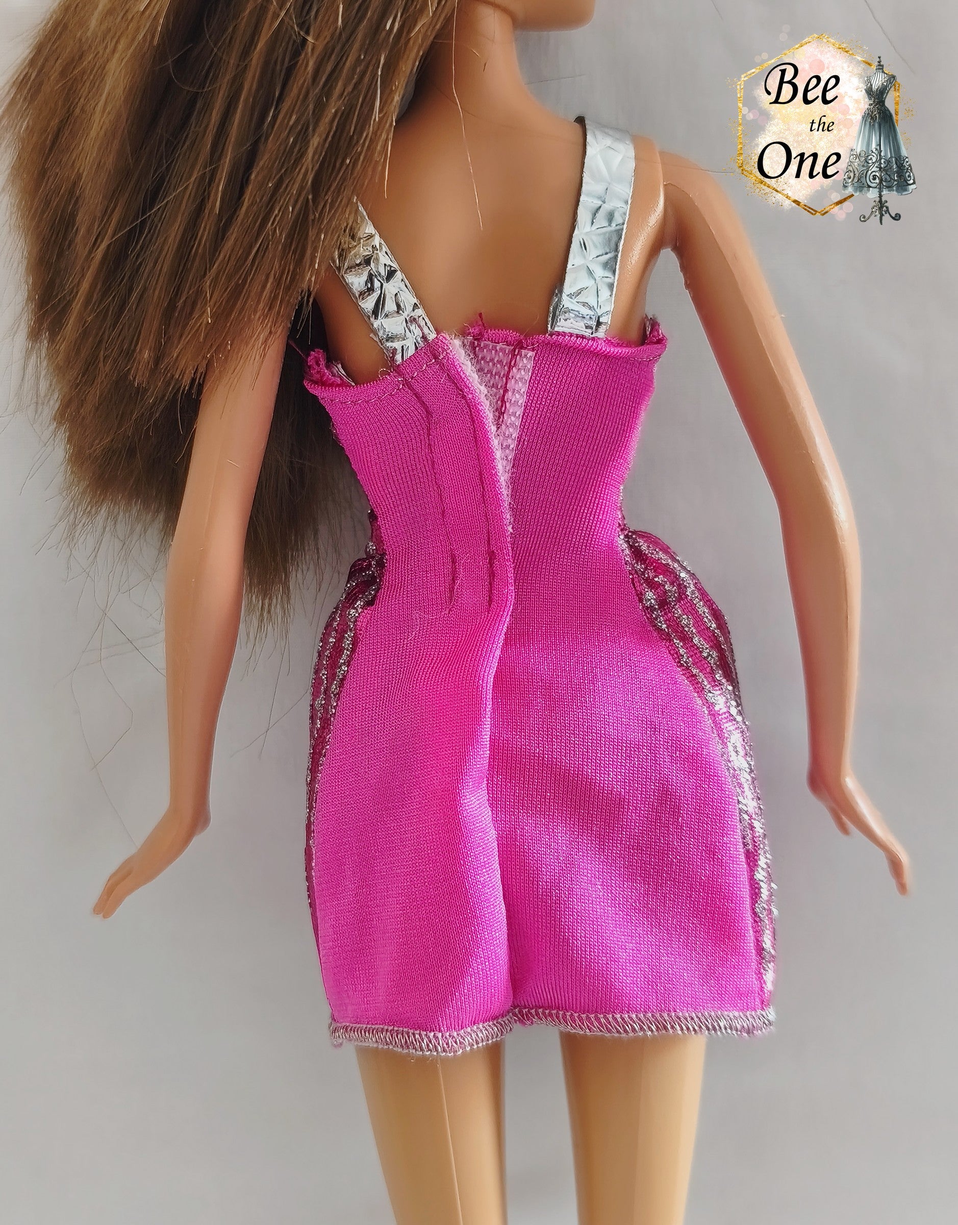 Robe courte de fête rose fuchsia brillante avec rayures verticales pailletées argentées, bretelles métallisées et découpes géométriques en losanges à la taille, vêtement d'origine pour poupée Barbie Fashionistas 2013, portée par une poupée brune sur fond blanc. Gros plan dos
