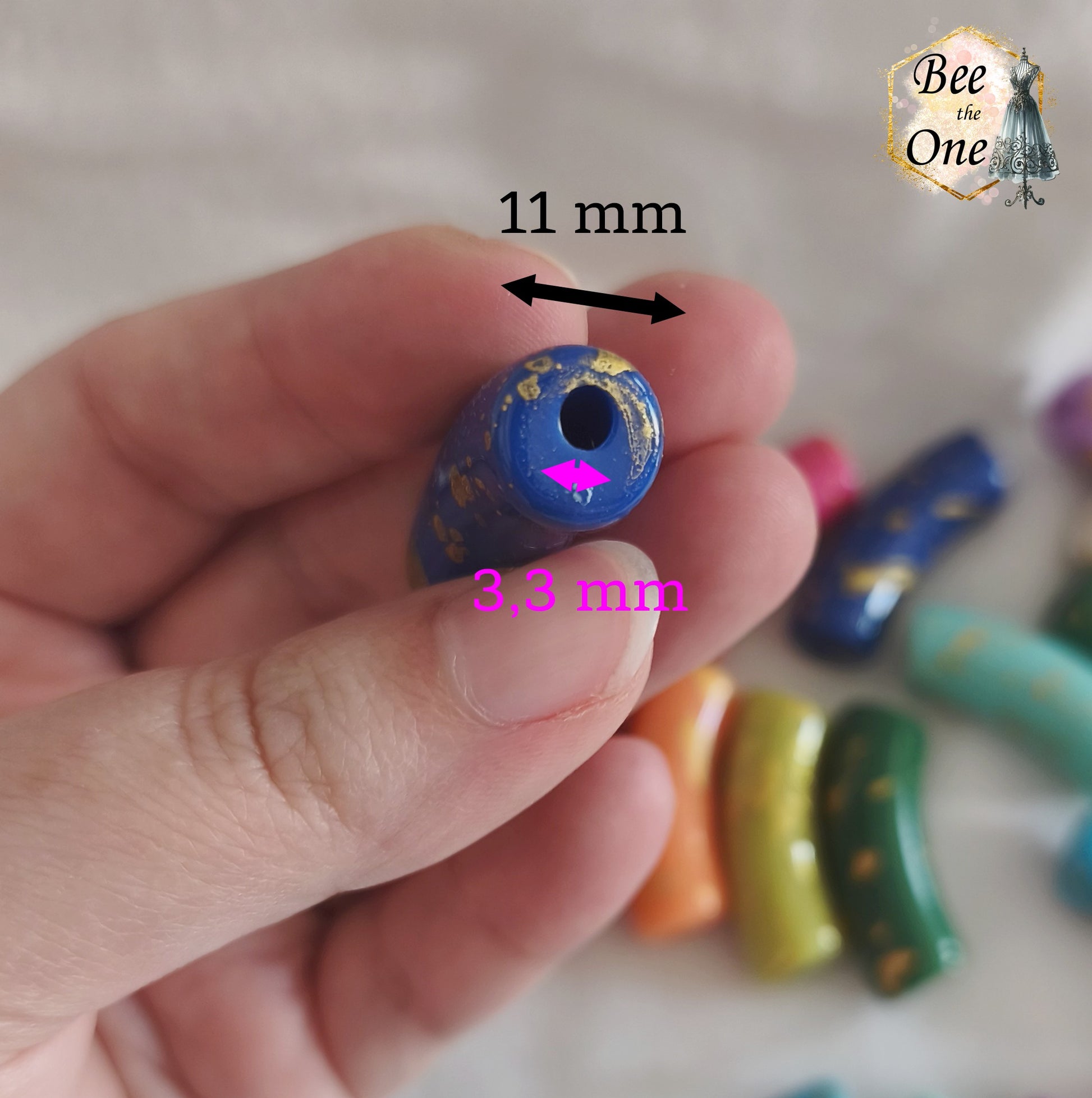 Assortiment de grandes perles tubes incurvées en acrylique mesurant 34x11 mm. Les perles sont multicolores (bleu, vert, rouge, rose, violet, orange, blanc) et ornées d'éclats et de mouchetures dorées, disposées sur un fond blanc. Gros plan trou
