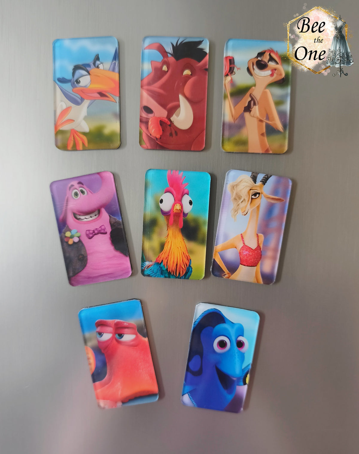 Série de huit magnets rectangulaires verticaux à bords arrondis représentant des personnages animés Disney et Pixar : Zazu, Pumbaa, Timon, Bing Bong, Hei Hei, Gazelle, Hank et Dory, disposés sur une surface métallique grise.