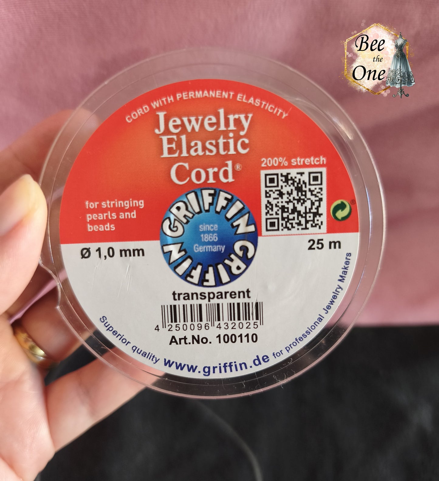 Bobine ronde de fil élastique transparent pour bijoux de la marque Griffin (Jewelry Elastic Cord) de diamètre 1,0 mm, avec un échantillon de fil déroulé sur un fond bicolore rose et noir. Gros plan sur l'étiquette de la bobine