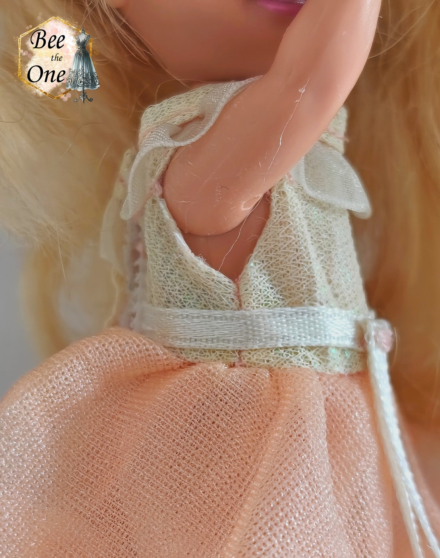 Petite robe de princesse pour poupée Kelly (Shelly), issue du film Barbie et le Cheval Magique (2005). Le haut est blanc scintillant avec un col volanté blanc, la jupe est en tulle pêche, portée par une petite poupée blonde sur fond blanc. gros plan côté droit