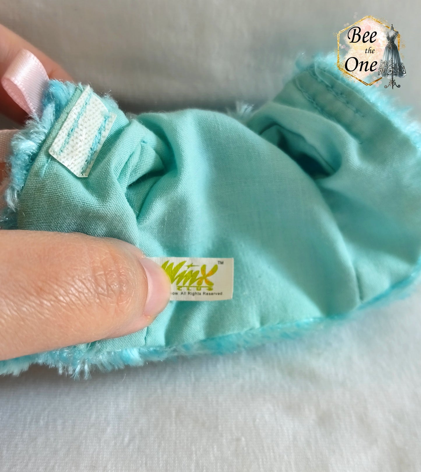 Manteau court en fausse fourrure bleu turquoise avec noeud en ruban rose satiné pour poupée Winx Club, issu du set Fairy Lounge de Mattel, porté par une poupée Flora sur fond blanc. Gros plan intérieur