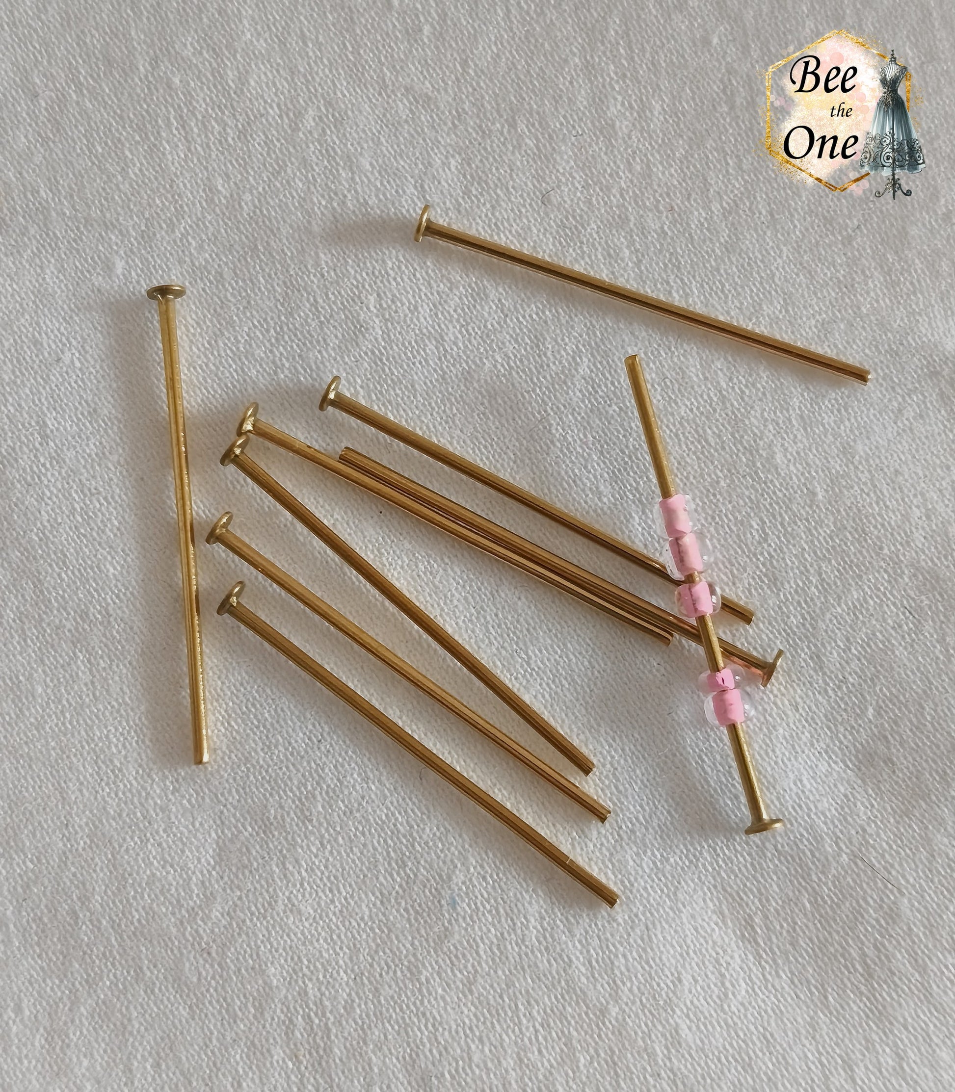 Clous de bijoutier à tête plate en acier inoxydable doré à l'or 18k de 30 mm, avec un clou montrant des petites perles de rocaille roses enfilées, présentés sur fond blanc texturé.