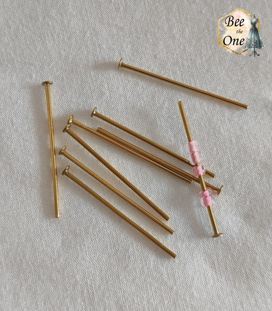 Clous de bijoutier à tête plate en acier inoxydable doré à l'or 18k de 30 mm, avec un clou montrant des petites perles de rocaille roses enfilées, présentés sur fond blanc texturé.