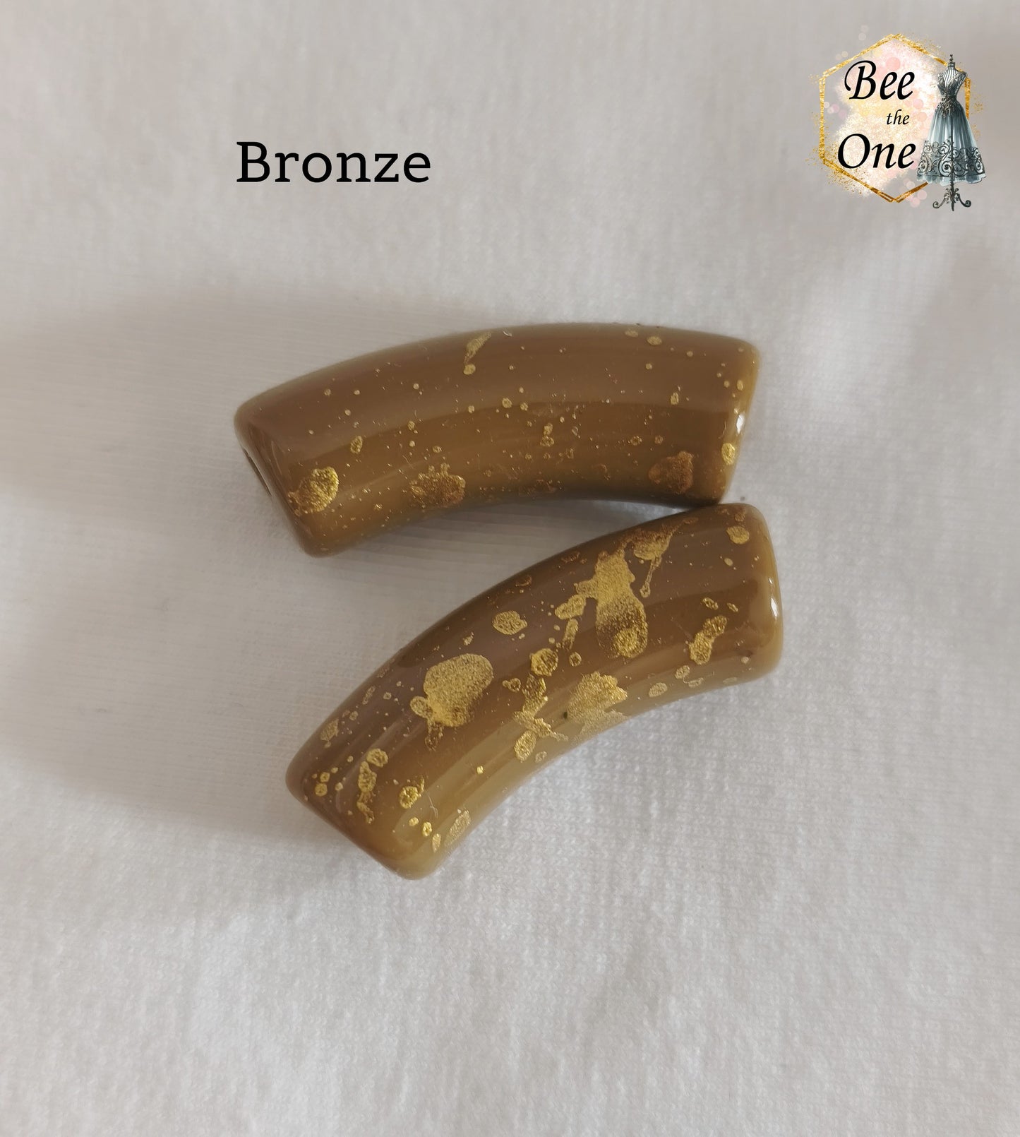 Grandes perles tubes incurvées en acrylique mesurant 34x11 mm. Les perles sont couleur bronze et ornées d'éclats et de mouchetures dorées, disposées sur un fond blanc.