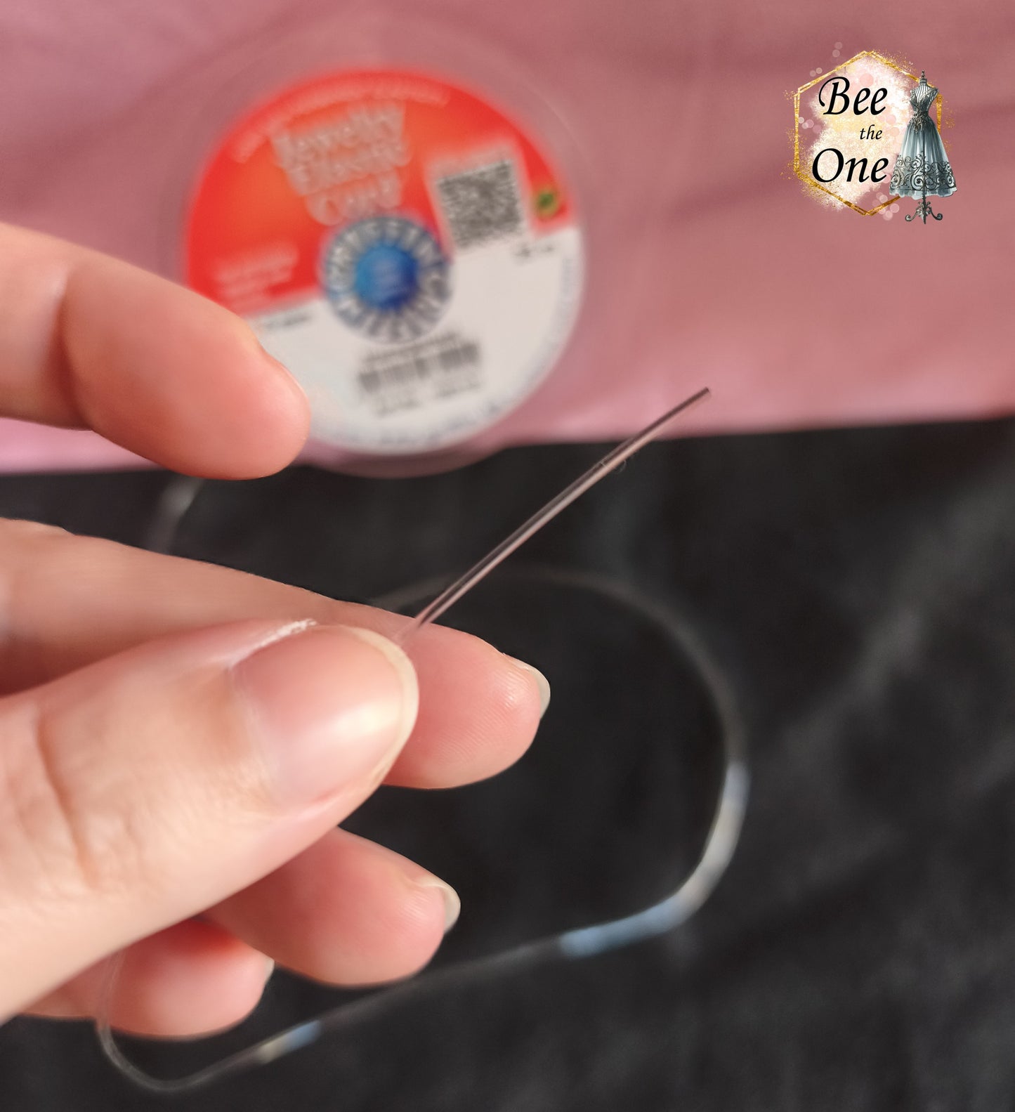 Bobine ronde de fil élastique transparent pour bijoux de la marque Griffin (Jewelry Elastic Cord) de diamètre 1,0 mm, avec un échantillon de fil déroulé sur un fond bicolore rose et noir. Gros plan sur le fil