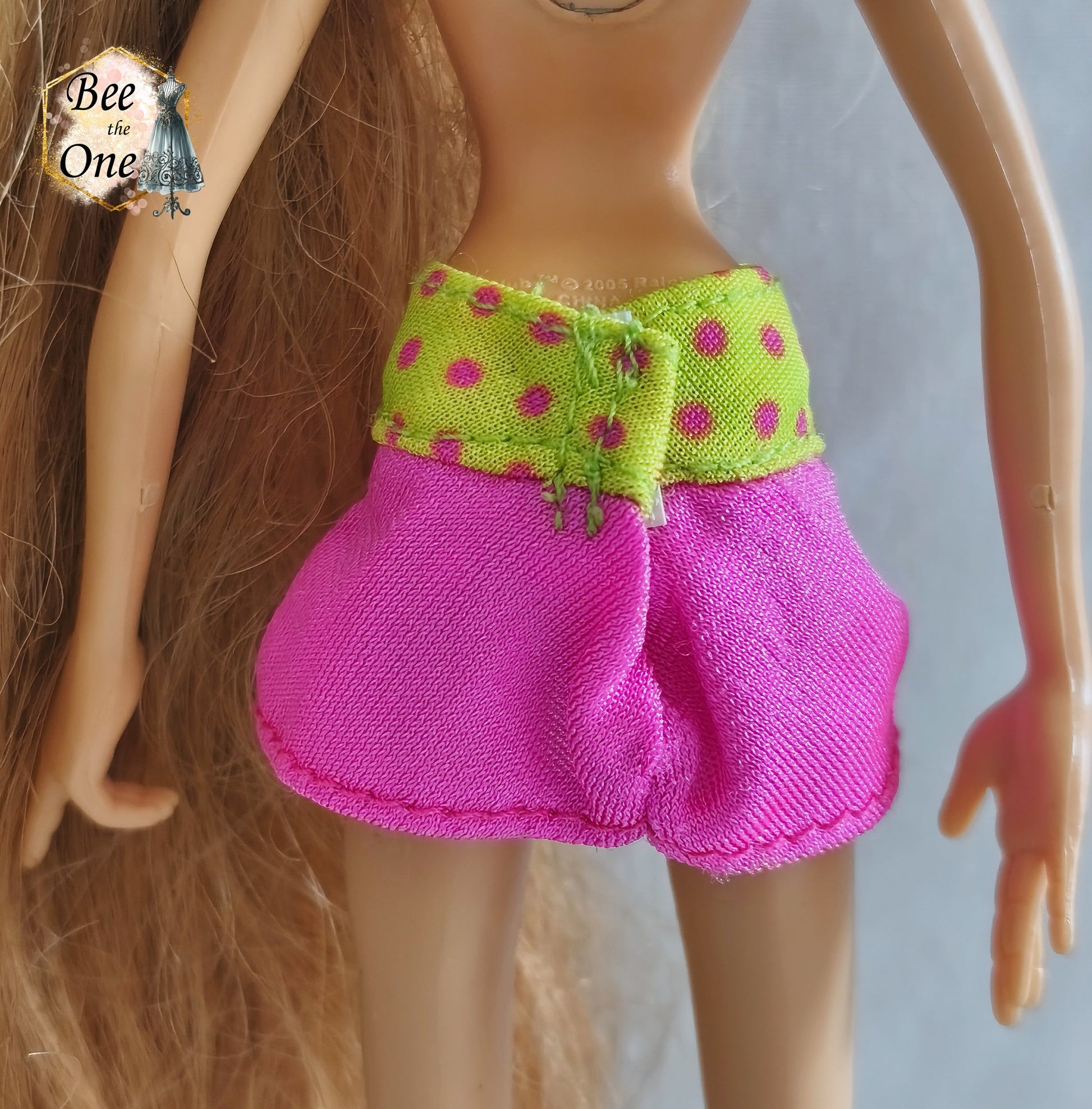 Mini-jupe rose avec large ceinture jaune fluo à pois roses pour poupée Winx Club, issue du modèle Flora Flutter Magic, portée par une poupée sur fond blanc. Gros plan dos