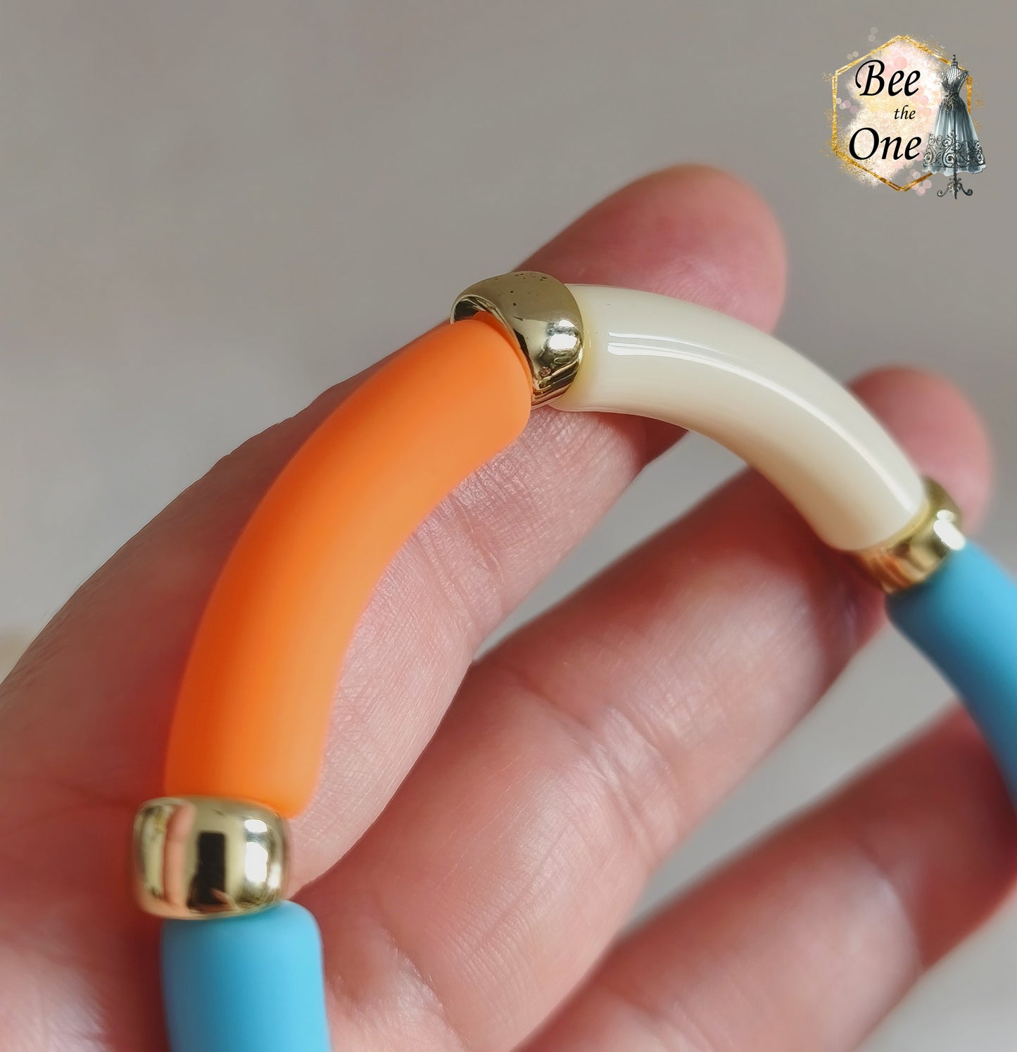Set de trois bracelets élastiques faits main pour adulte, composés de perles tubes incurvées en acrylique aux teintes bleu azur, orange vif et crème, avec détails marbrés dorés et séparateurs dorés. Gros plan sur une perle orange mat en acrylique