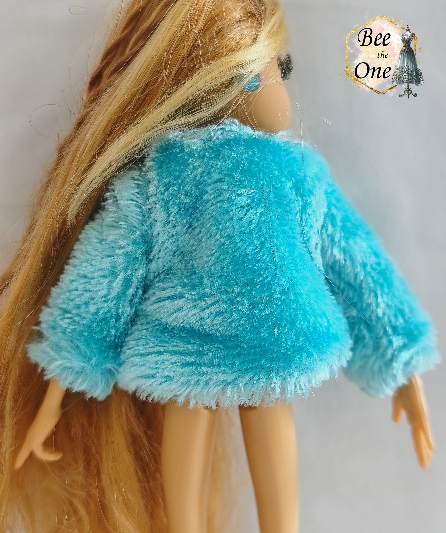 Manteau court en fausse fourrure bleu turquoise avec noeud en ruban rose satiné pour poupée Winx Club, issu du set Fairy Lounge de Mattel, porté par une poupée Flora sur fond blanc. Gros plan dos