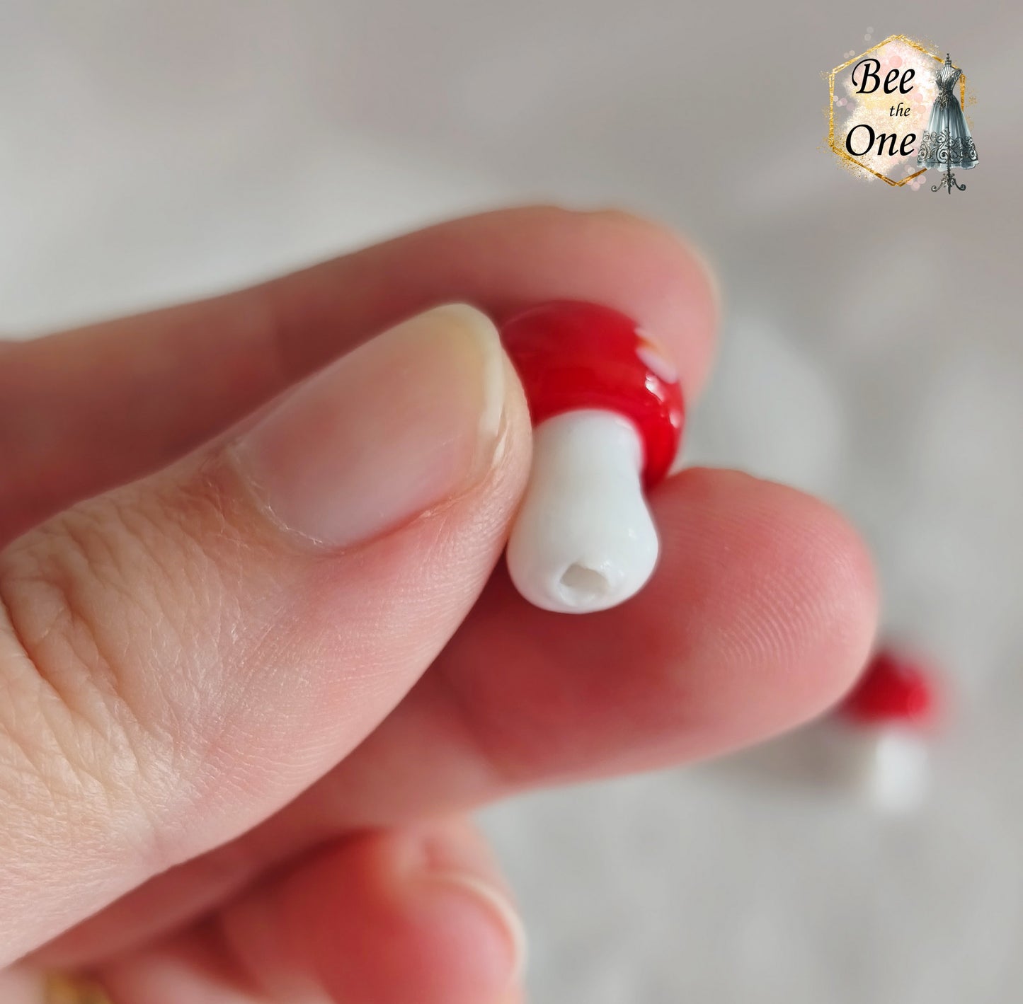 Perles en verre filé rouge et blanc en forme de champignon amanite tue-mouche à pois, percées verticalement, sur fond blanc. Gros plan trou du bas