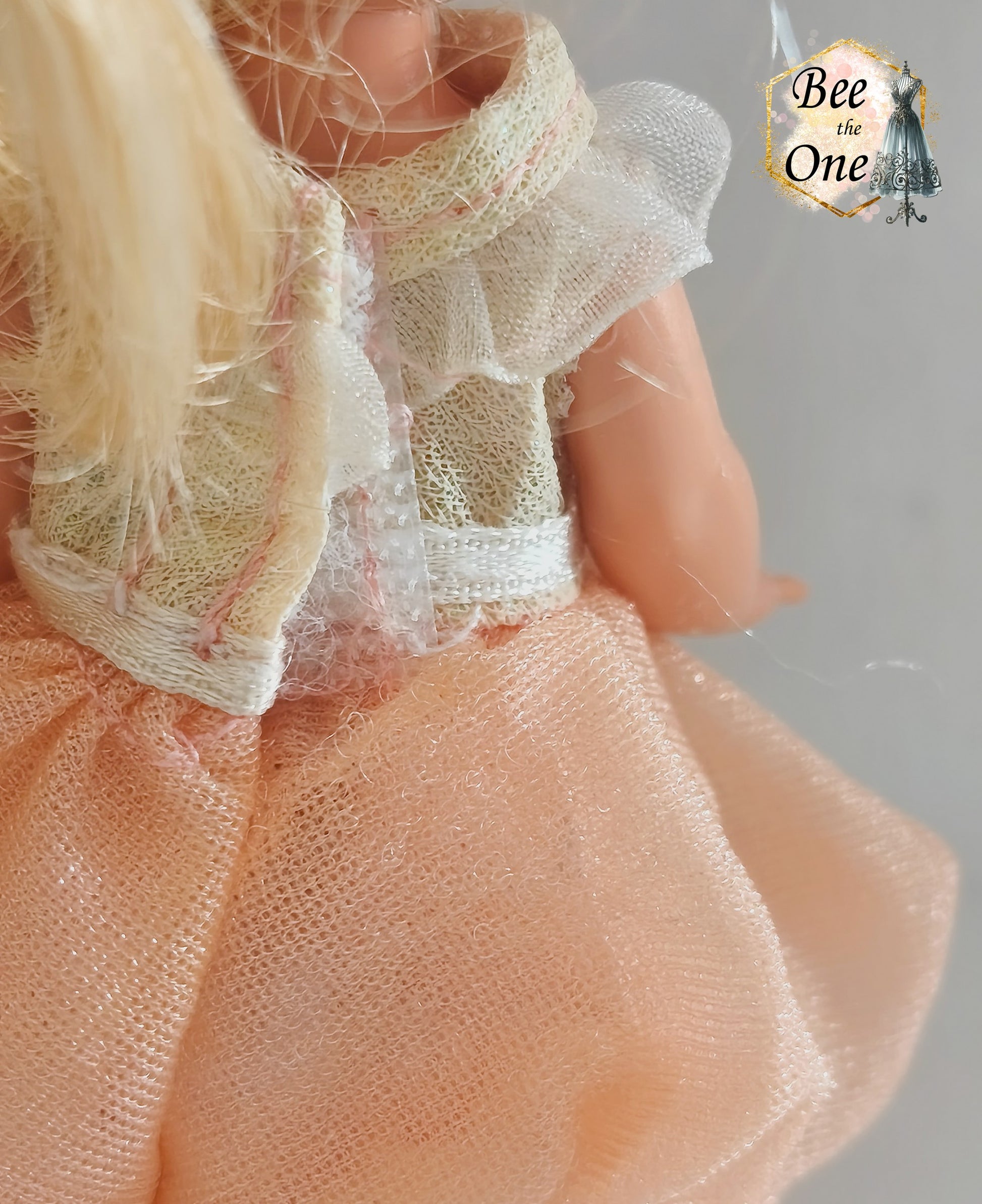 Petite robe de princesse pour poupée Kelly (Shelly), issue du film Barbie et le Cheval Magique (2005). Le haut est blanc scintillant avec un col volanté blanc, la jupe est en tulle pêche, portée par une petite poupée blonde sur fond blanc. Gros plan détail dos