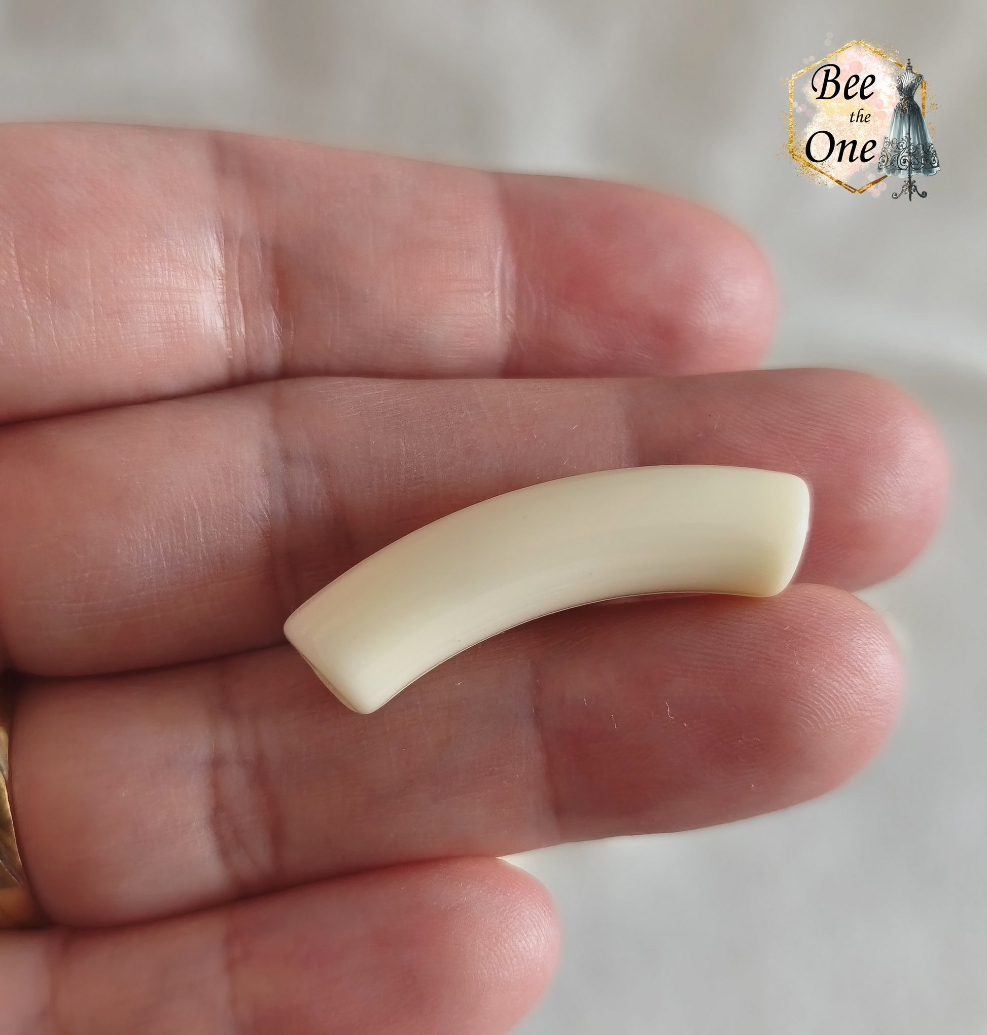 Perles tubes incurvées en acrylique couleur blanc crème unie mesurant 32x7 mm, disposées en demi-cercle sur un fond blanc texturé. Gros plan sur une perle