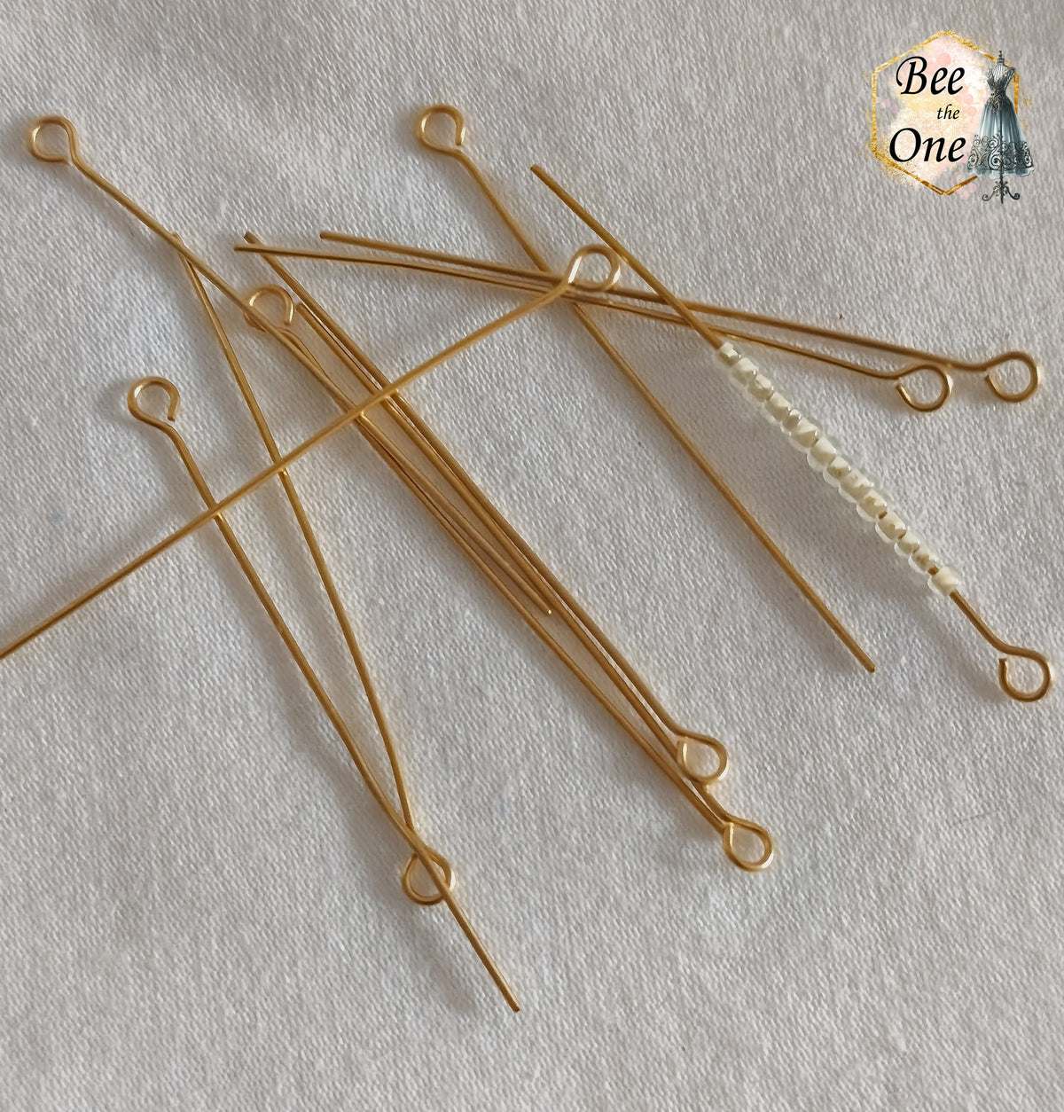 Lot de longs clous de bijoutier à œillet en acier inoxydable doré de 50 mm, avec un clou montrant une rangée de perles de rocaille ivoire enfilées, sur fond blanc texturé.