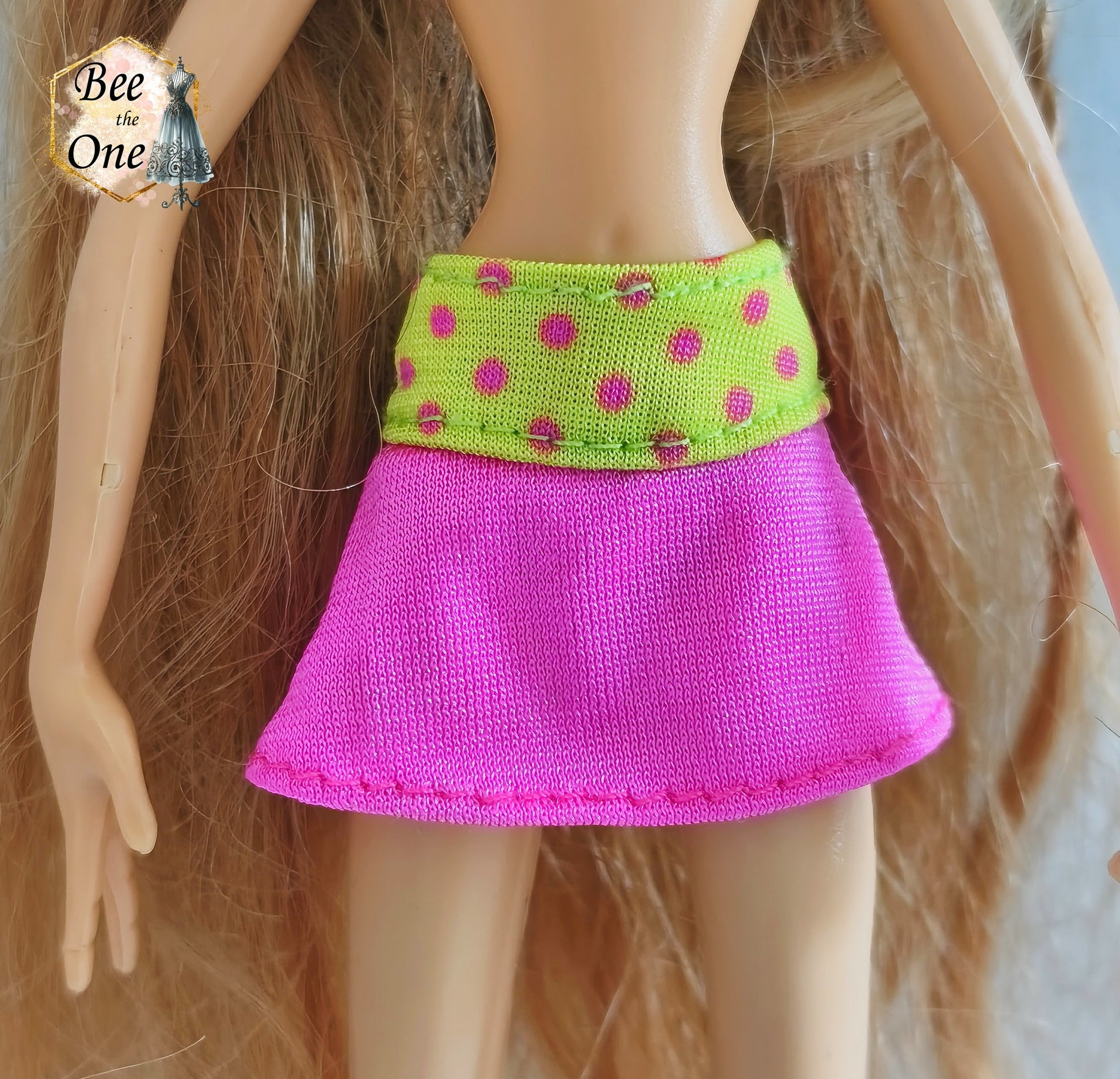 Mini-jupe rose avec large ceinture jaune fluo à pois roses pour poupée Winx Club, issue du modèle Flora Flutter Magic, portée par une poupée sur fond blanc. Gros plan devant