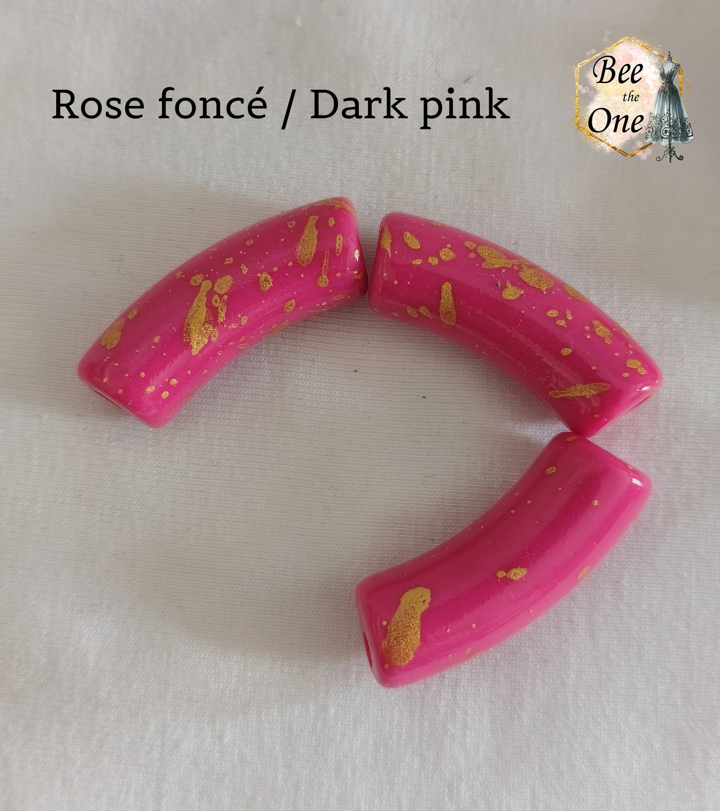 Grandes perles tubes incurvées en acrylique mesurant 34x11 mm. Les perles sont couleur rose foncé et ornées d'éclats et de mouchetures dorées, disposées sur un fond blanc.