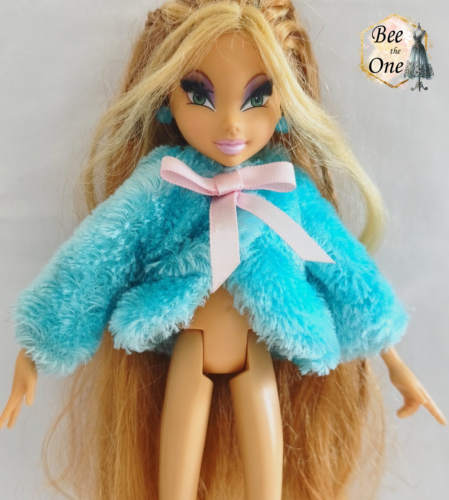 Manteau court en fausse fourrure bleu turquoise avec noeud en ruban rose satiné pour poupée Winx Club, issu du set Fairy Lounge de Mattel, porté par une poupée Flora sur fond blanc. Gros plan devant