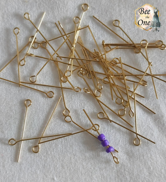 Lot de clous de bijoutier à œillet en acier inoxydable doré à l'or 18k de 26 mm, avec un clou montrant trois perles violettes enfilées, présentés sur fond blanc texturé.