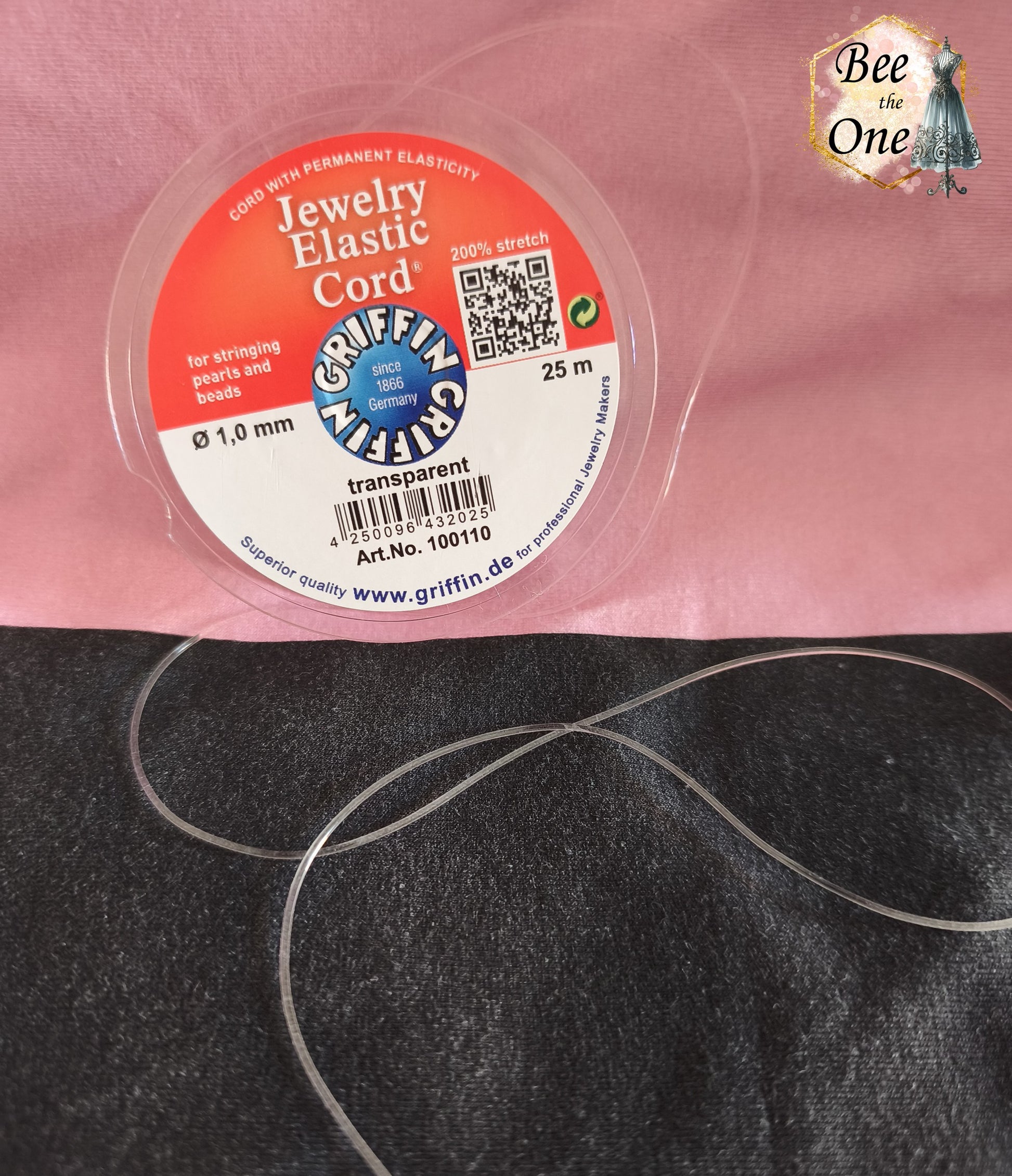 Bobine ronde de fil élastique transparent pour bijoux de la marque Griffin (Jewelry Elastic Cord) de diamètre 1,0 mm, avec un échantillon de fil déroulé sur un fond bicolore rose et noir.