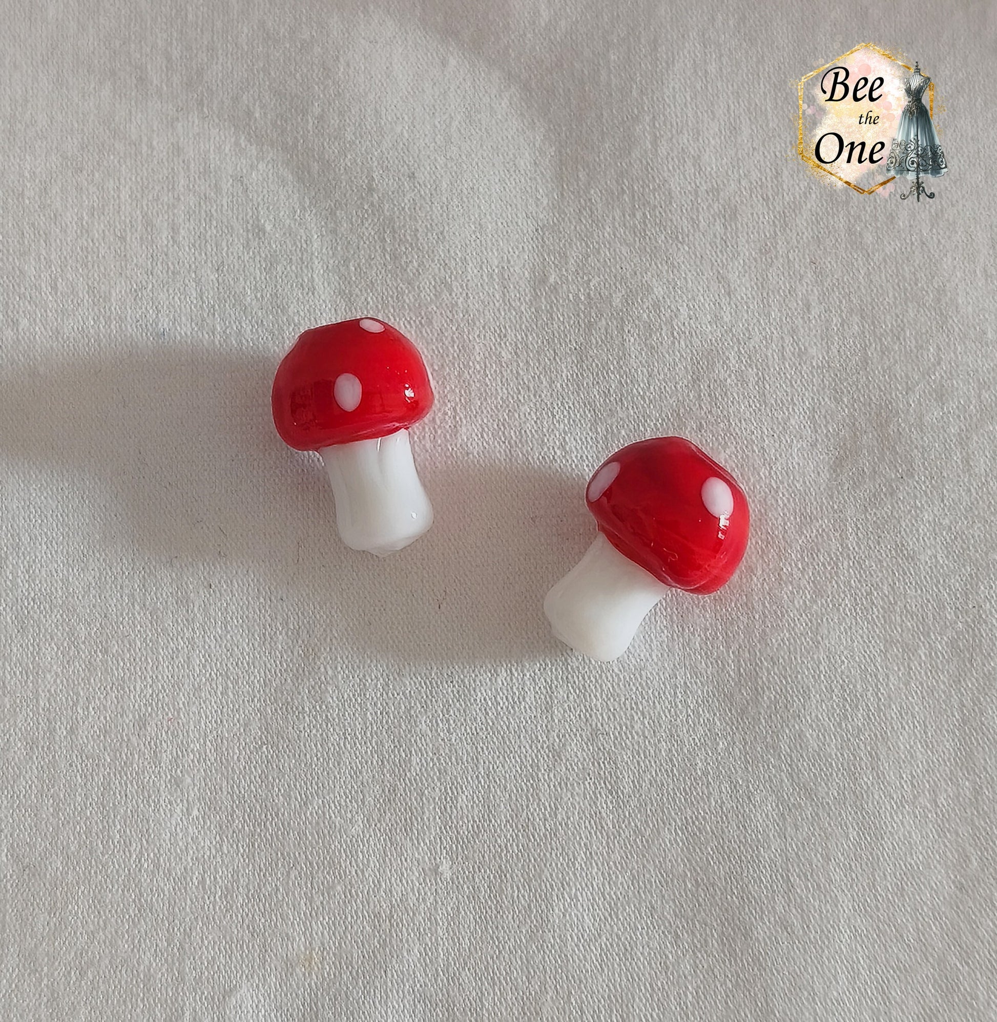 Perles en verre filé rouge et blanc en forme de champignon amanite tue-mouche à pois, percées verticalement, sur fond blanc.