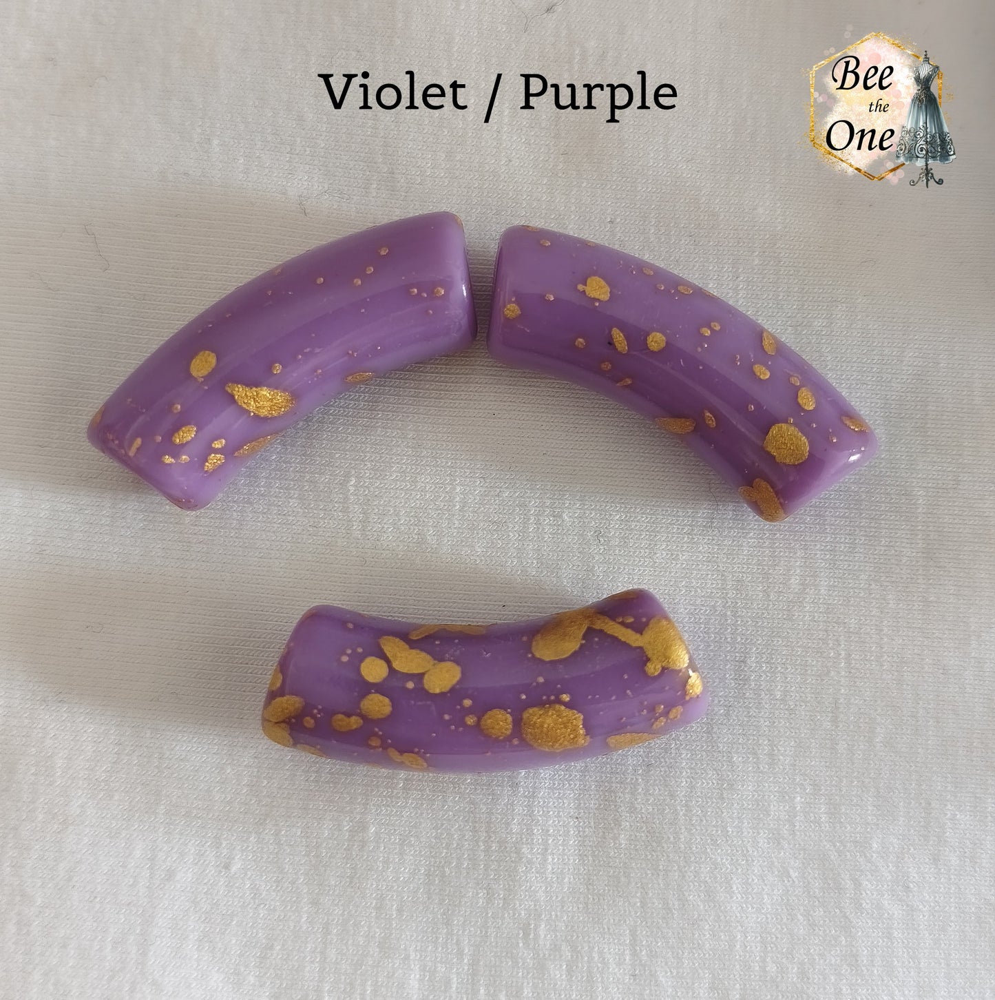 Grandes perles tubes incurvées en acrylique mesurant 34x11 mm. Les perles sont couleur violet et ornées d'éclats et de mouchetures dorées, disposées sur un fond blanc.
