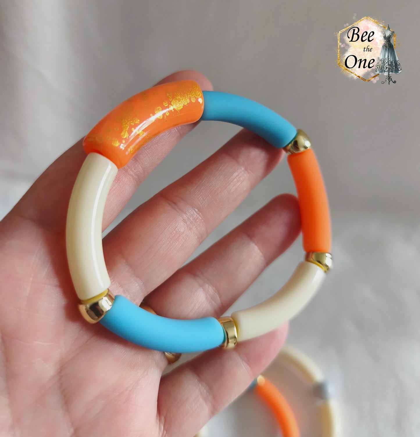 Set de trois bracelets élastiques faits main pour adulte, composés de perles tubes incurvées en acrylique aux teintes bleu azur, orange vif et crème, avec détails marbrés dorés et séparateurs dorés. Gros plan sur une perle orange à marbrures dorées en acrylique