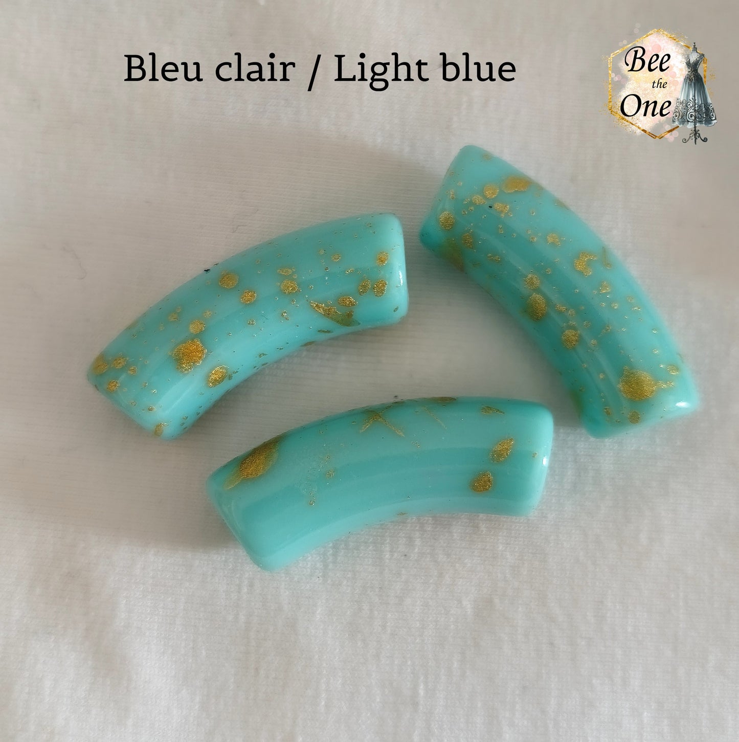 Grandes perles tubes incurvées en acrylique mesurant 34x11 mm. Les perles sont couleur bleu clair et ornées d'éclats et de mouchetures dorées, disposées sur un fond blanc.