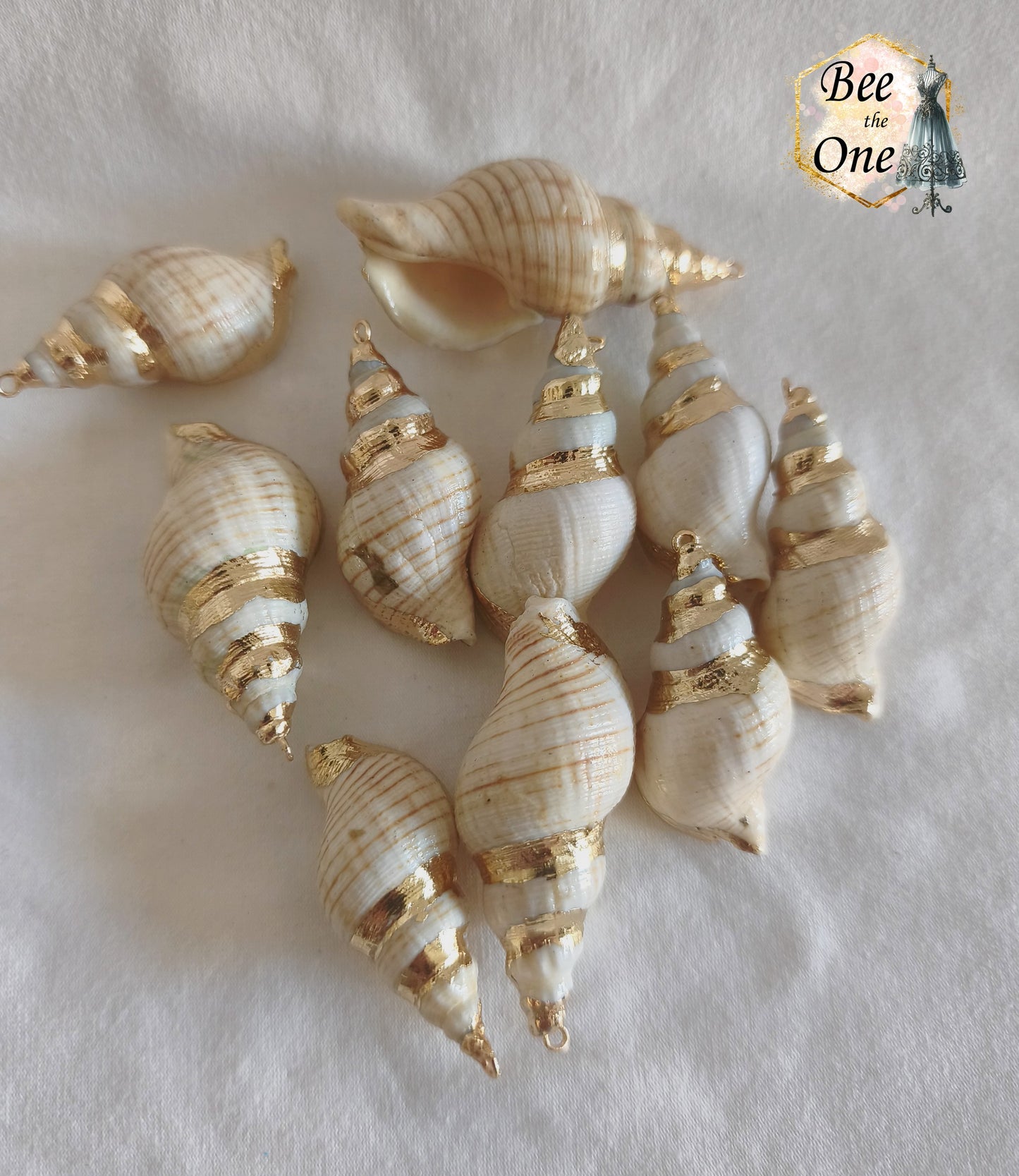 Grands pendentifs en coquillage naturel buccin (bulot) de 41 à 59 mm, orné d'une dorure en spirale au laiton qui suit les stries naturelles et muni d'un anneau d'attache, posés sur un fond blanc texturé.