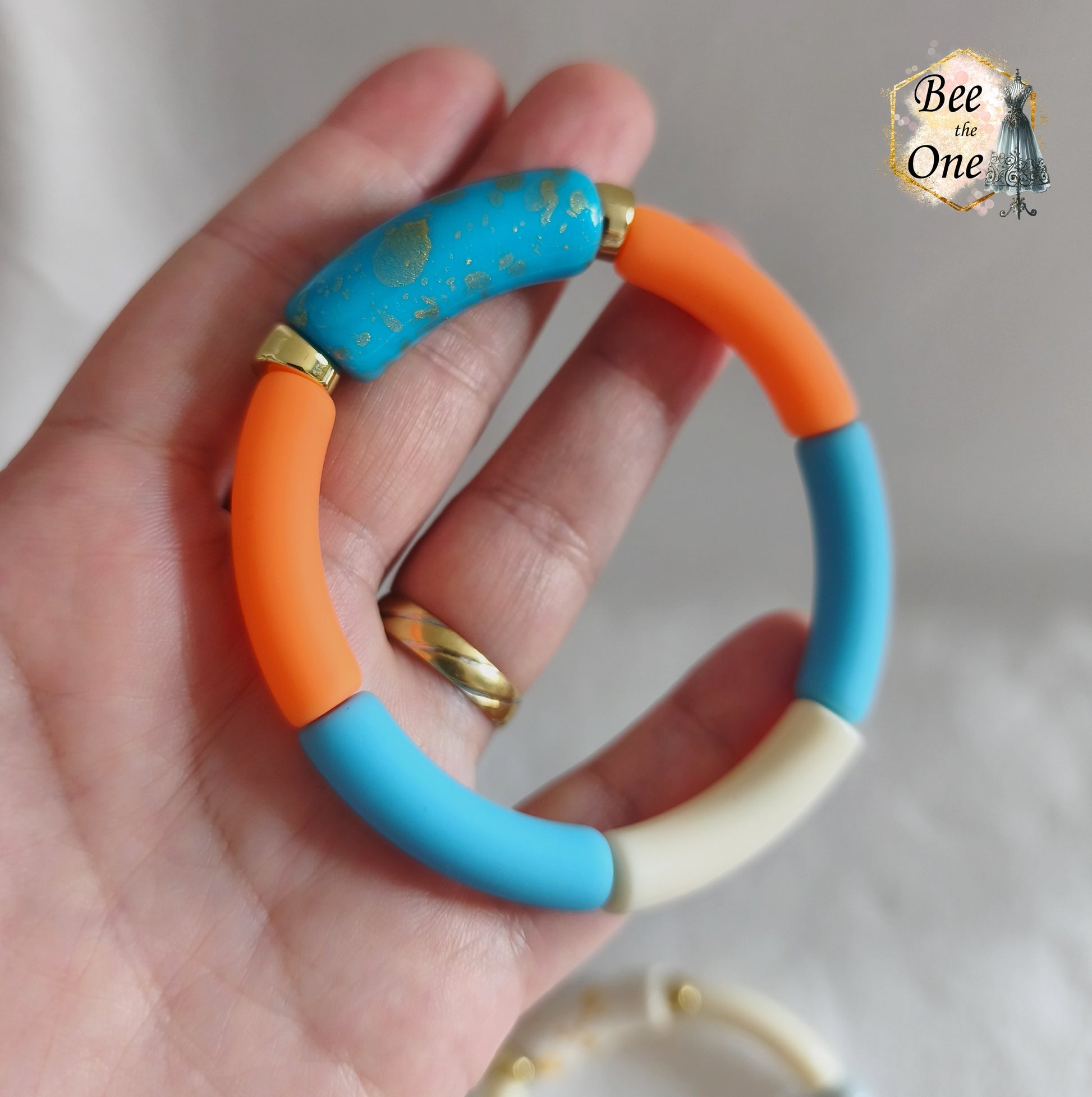 Set de trois bracelets élastiques faits main pour adulte, composés de perles tubes incurvées en acrylique aux teintes bleu azur, orange vif et crème, avec détails marbrés dorés et séparateurs dorés. Gros plan sur une perle bleue à marbrures dorées en acrylique