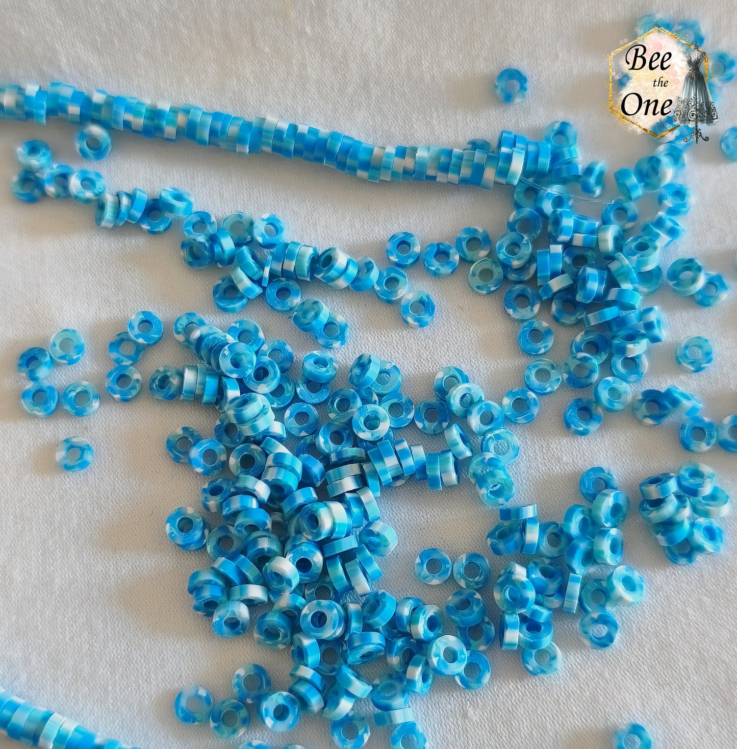 Petites perles rondelles Heishi Katsuki de 4 mm en pâte polymère, bleues, présentées sur tissu blanc.
